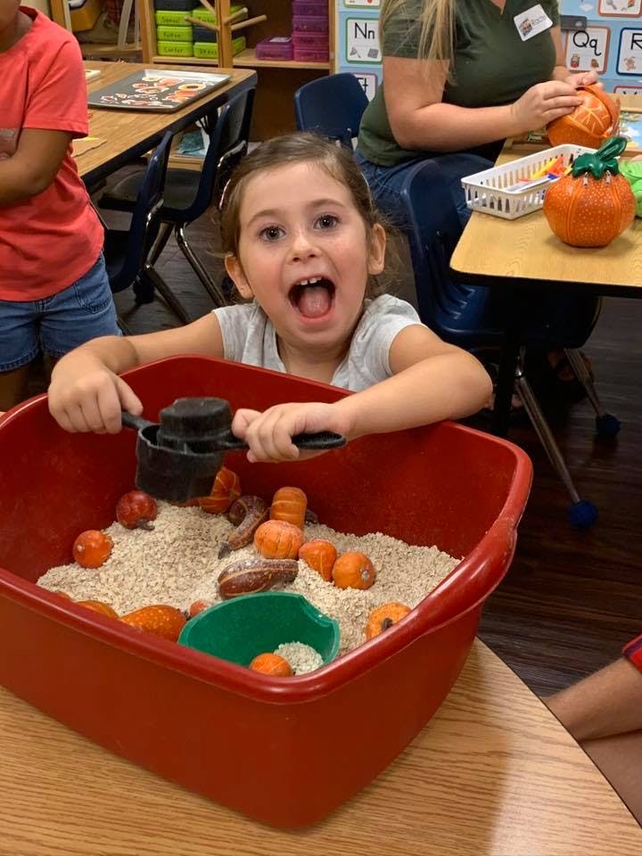 Child playing with a scoop in a red bin filled with grain and small pumpkins