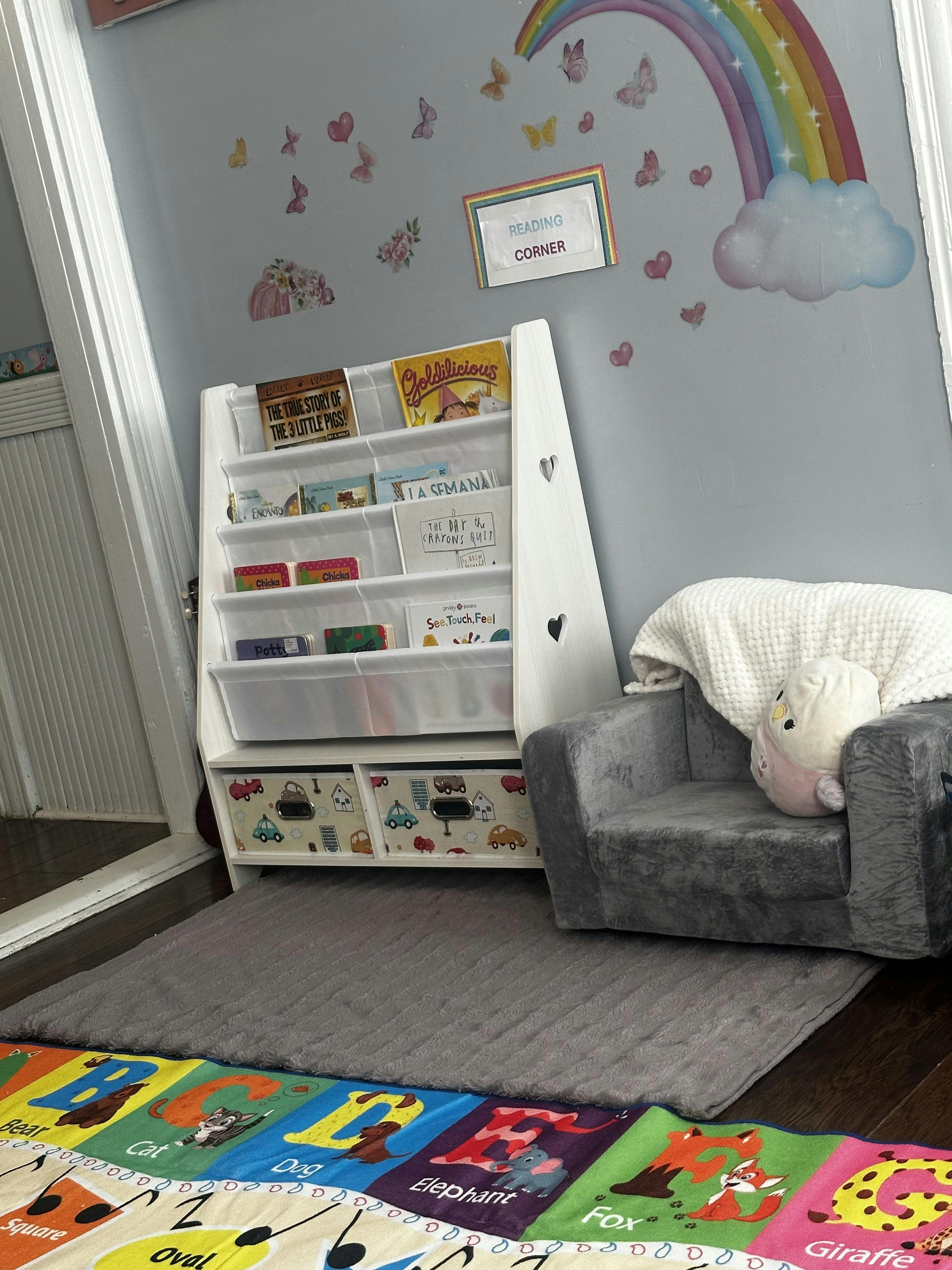 A children's reading corner with a bookshelf, gray chair, and alphabet mat