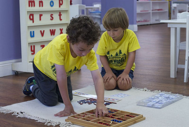 Two children kneeling on a rug, one playing with a wooden sorting tray