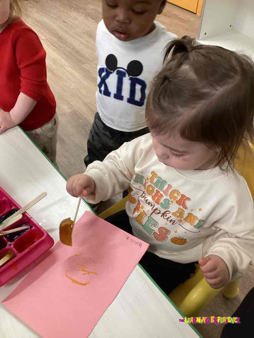 Child painting on pink paper with another child watching in background
