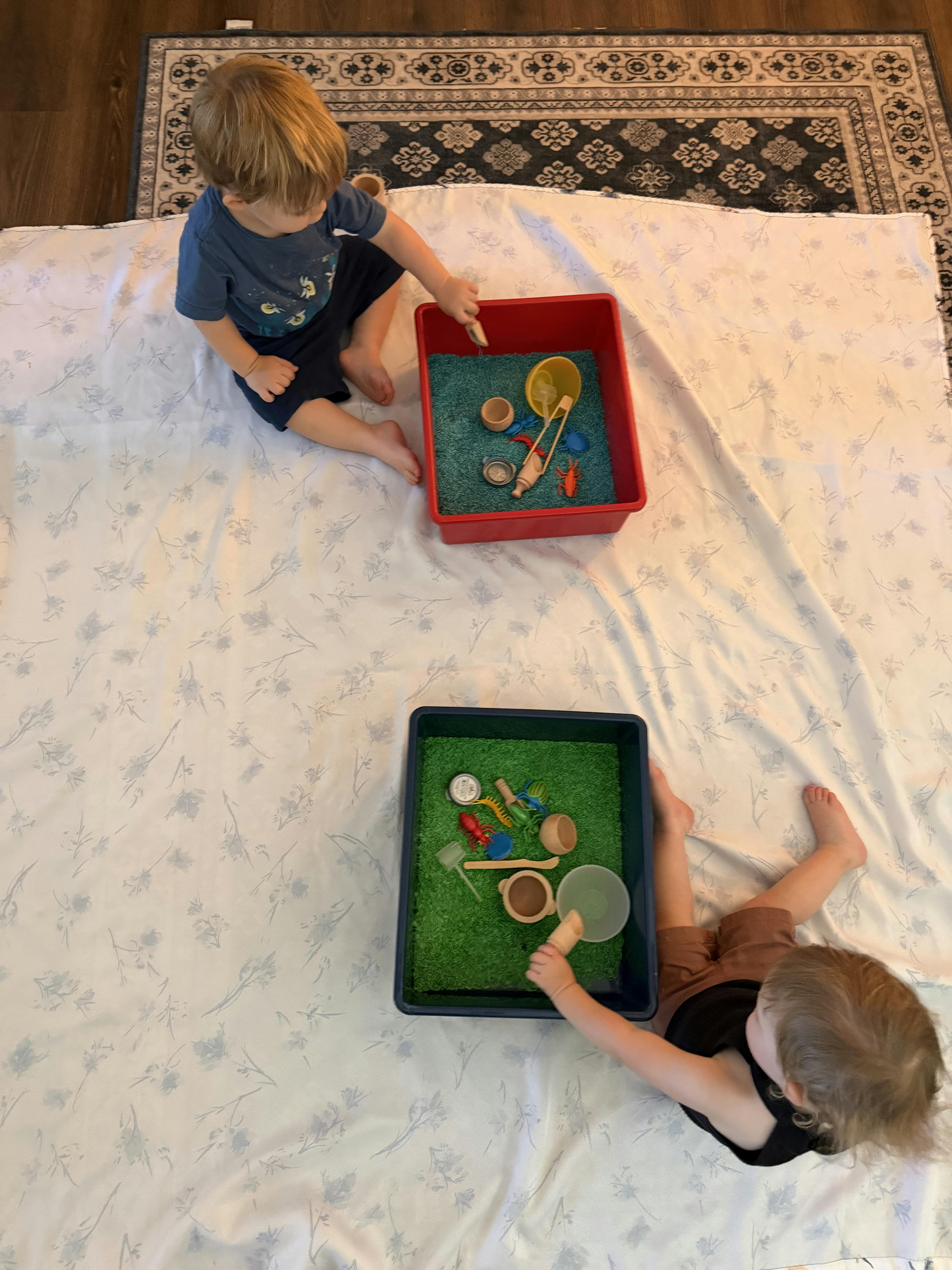 Two children playing with small toys in sensory bins on a blanket