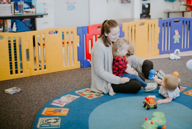 Person sitting on floor with two children playing with toys in a classroom