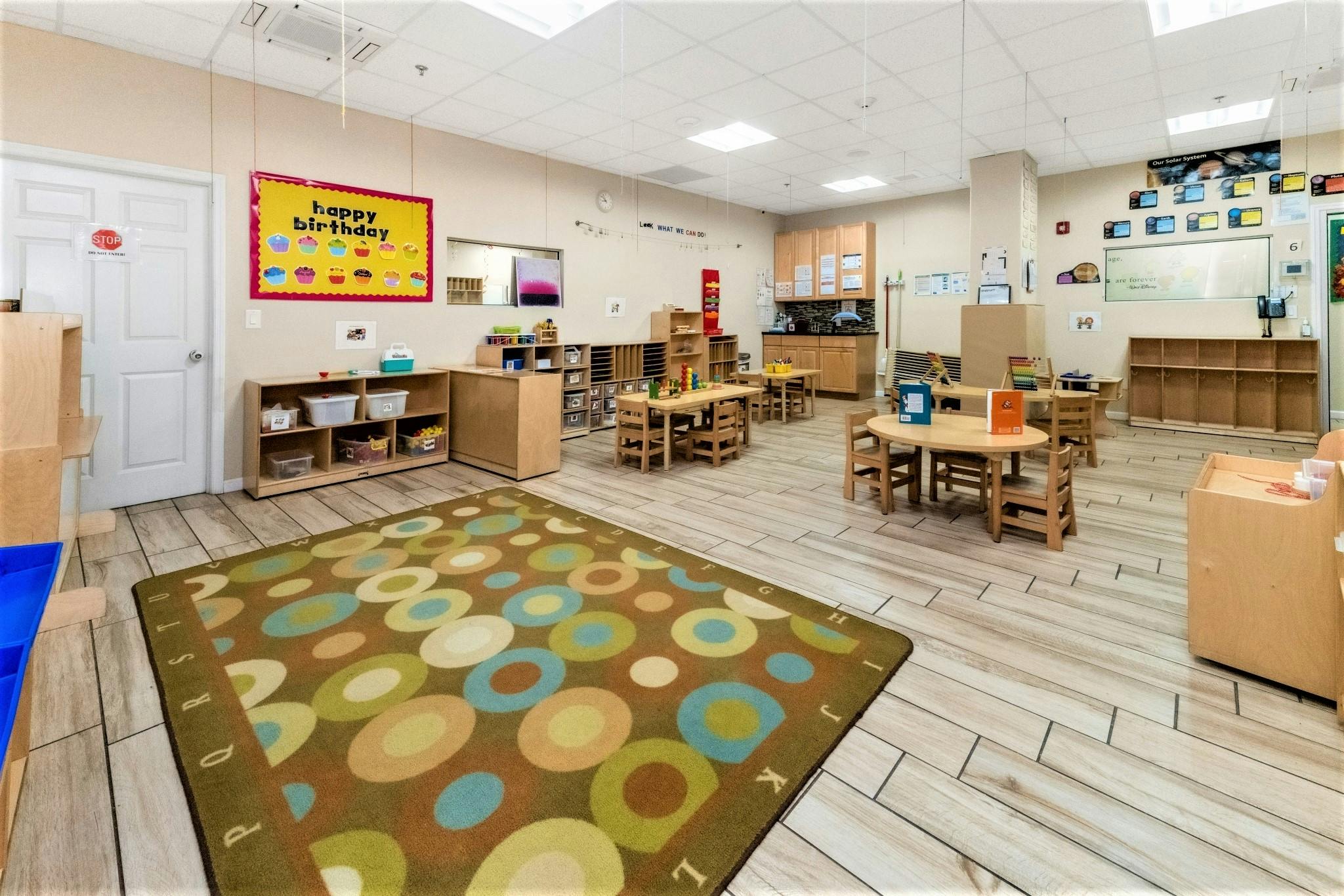 Empty classroom with small tables and chairs, a rug, and shelves