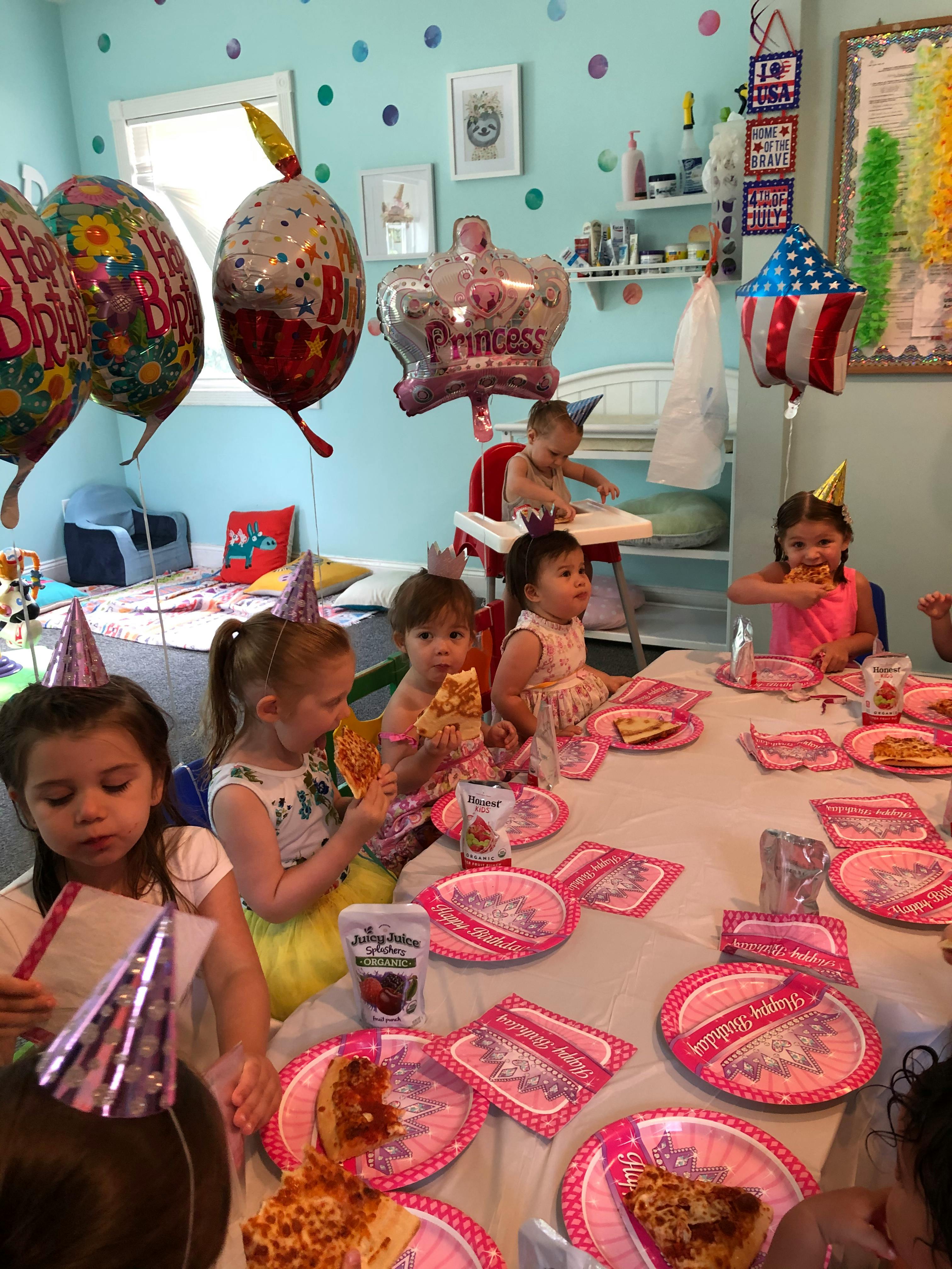 Children wearing party hats eating pizza at a table with balloons