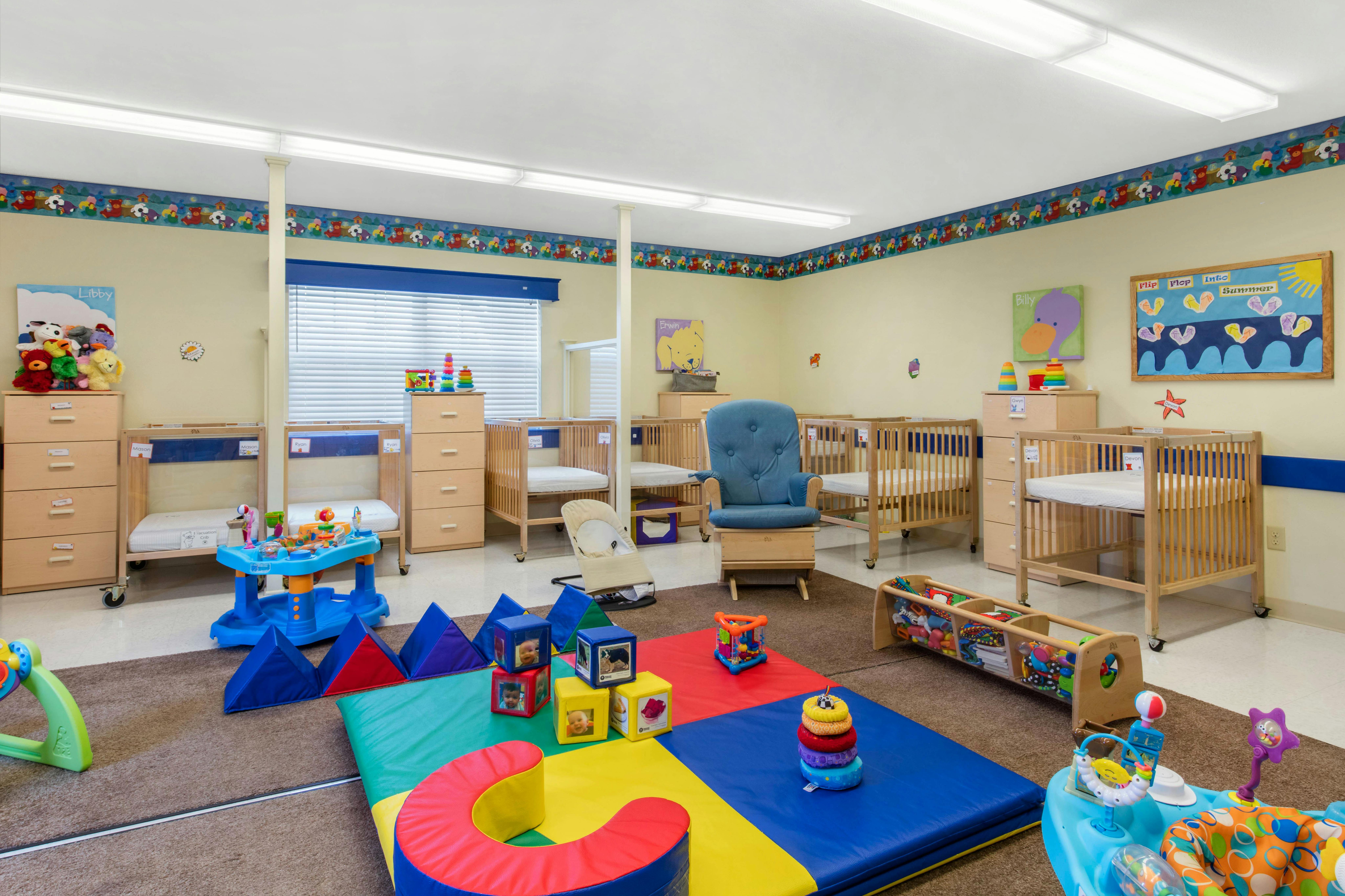 Baby room with multiple cribs, a blue rocking chair, and colorful play mats on the floor