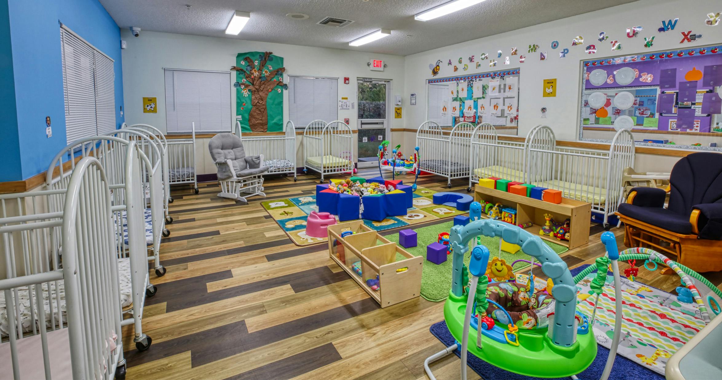 A colorful daycare room with several white cribs, a play mat, and toys