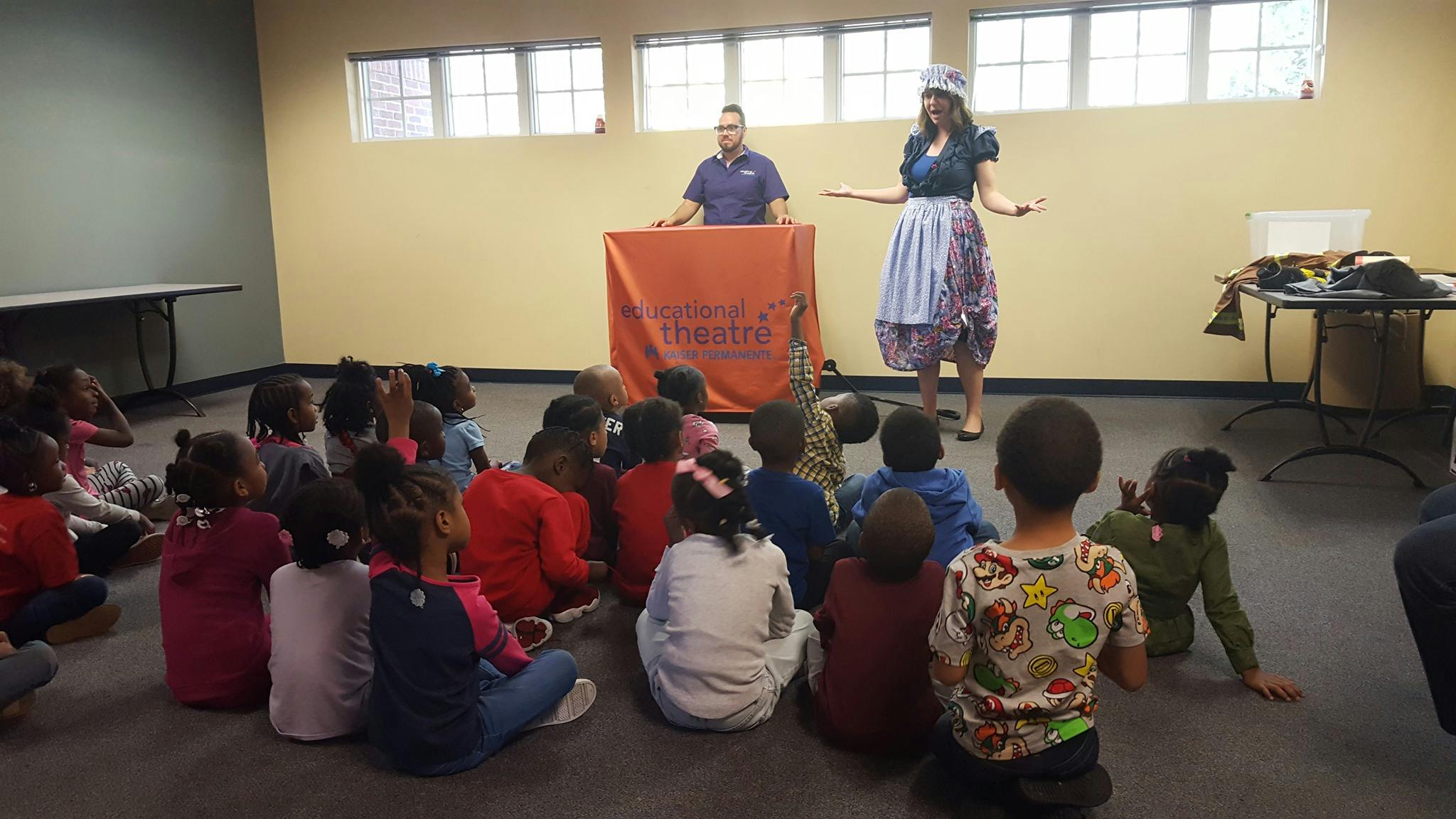 Children sitting on the floor watching two adults standing at the front of a room