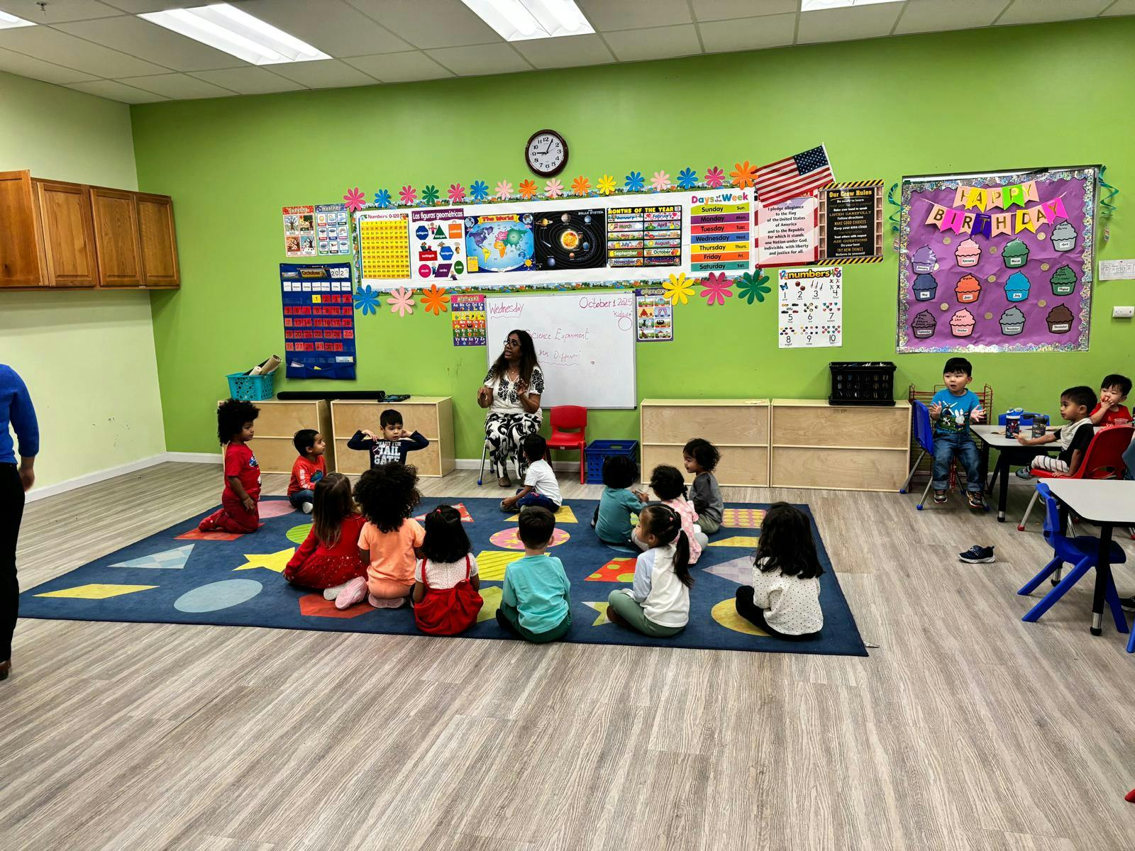 People in a classroom, with children sitting on a mat facing a person at a whiteboard