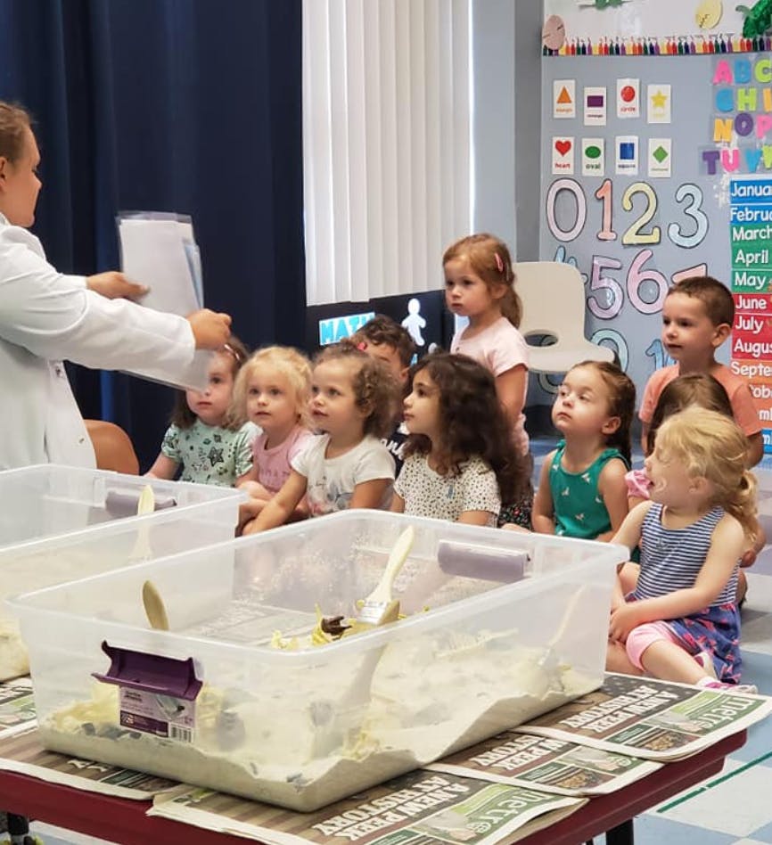 Person in lab coat showing papers to children sitting at a table with sensory bins