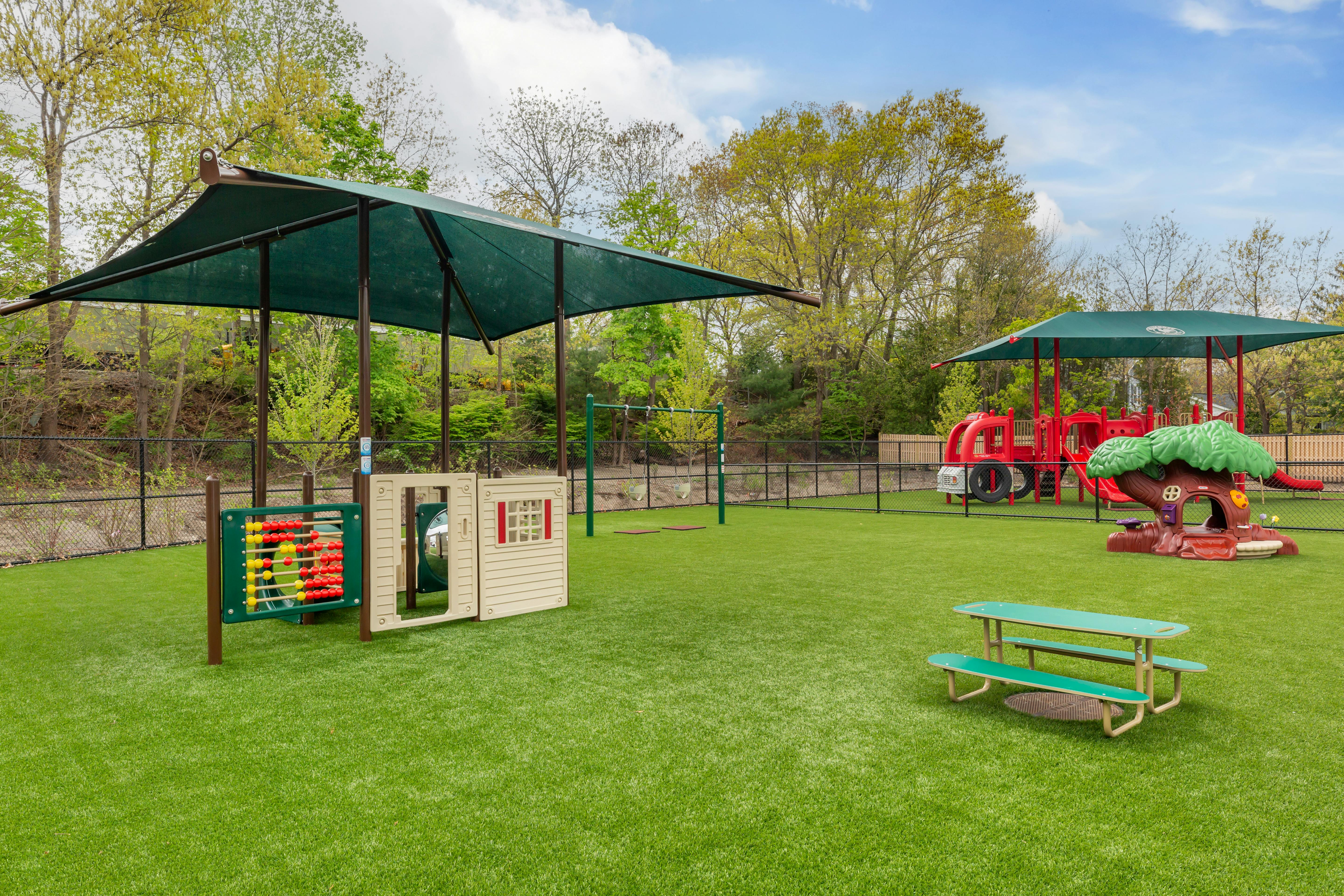 Outdoor playground with green turf, play structures, swings, and shade coverings