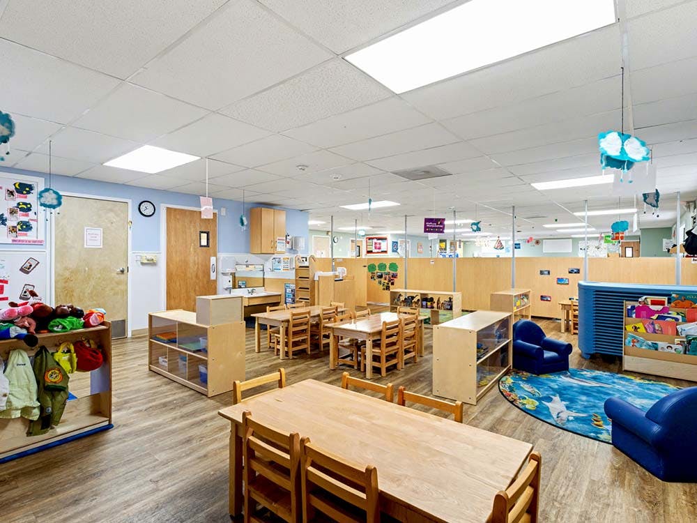 Classroom with wooden tables and chairs, play kitchen, and blue rug with armchairs