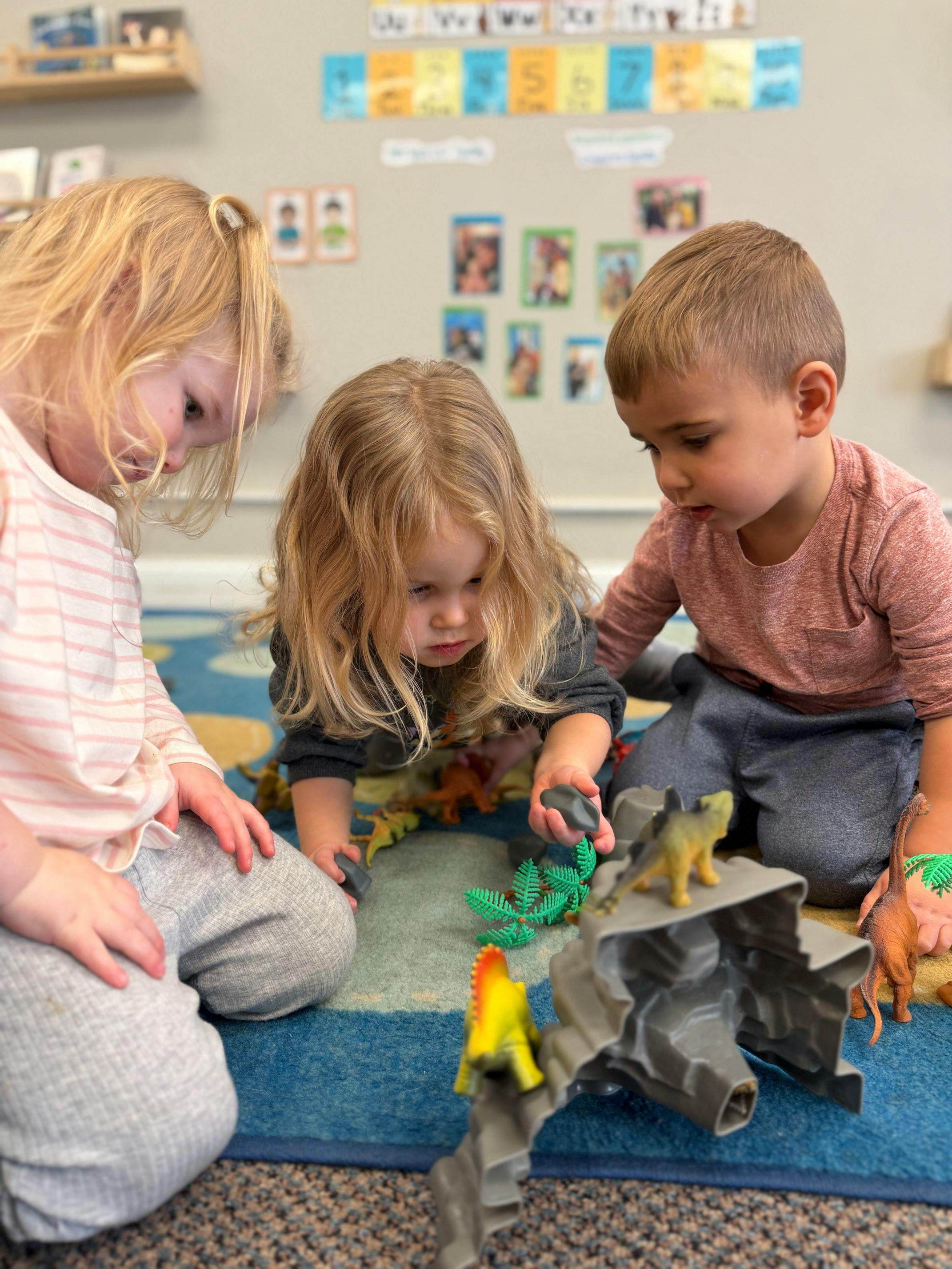 Three children playing with dinosaur toys in a classroom