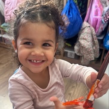Child smiling and holding a paintbrush with orange paint on their hand