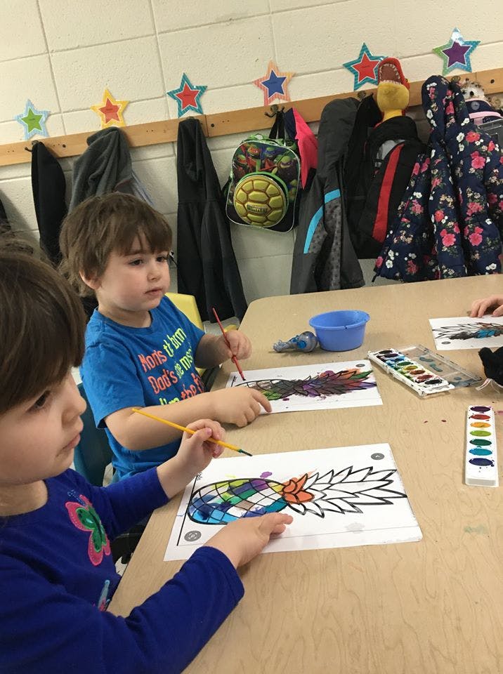 Two children painting pineapple outlines at a table in a classroom