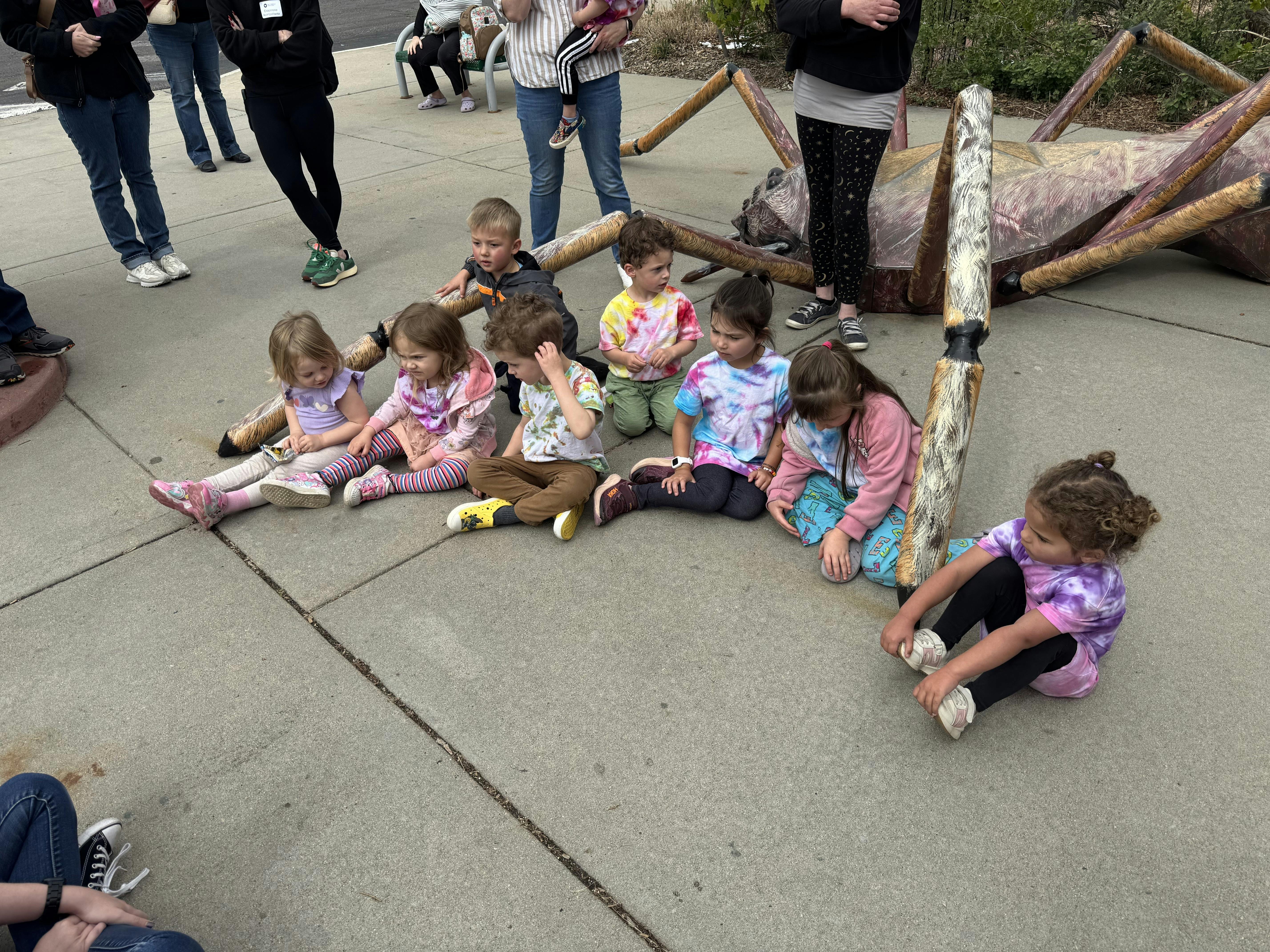 Group of children sitting on concrete next to large insect sculpture