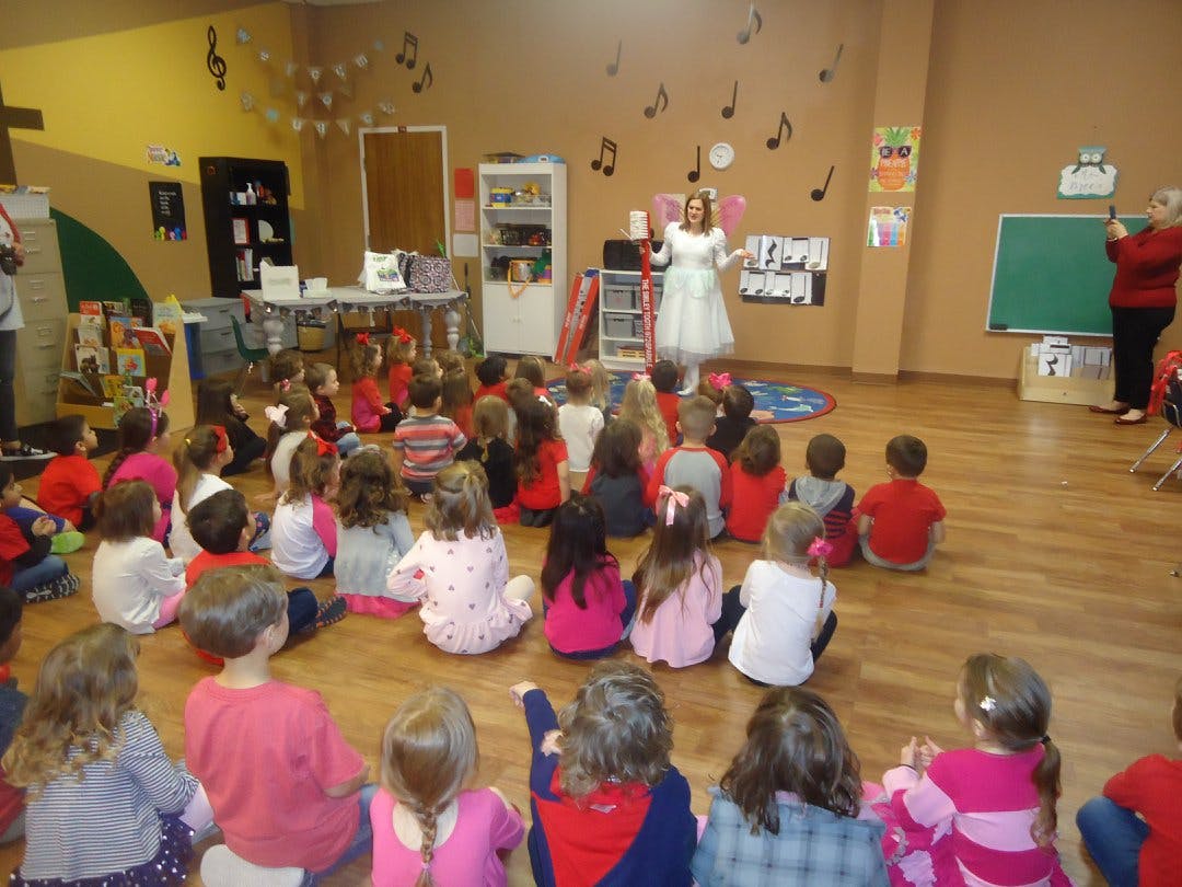 Person in white dress speaking to children sitting on the floor in a classroom