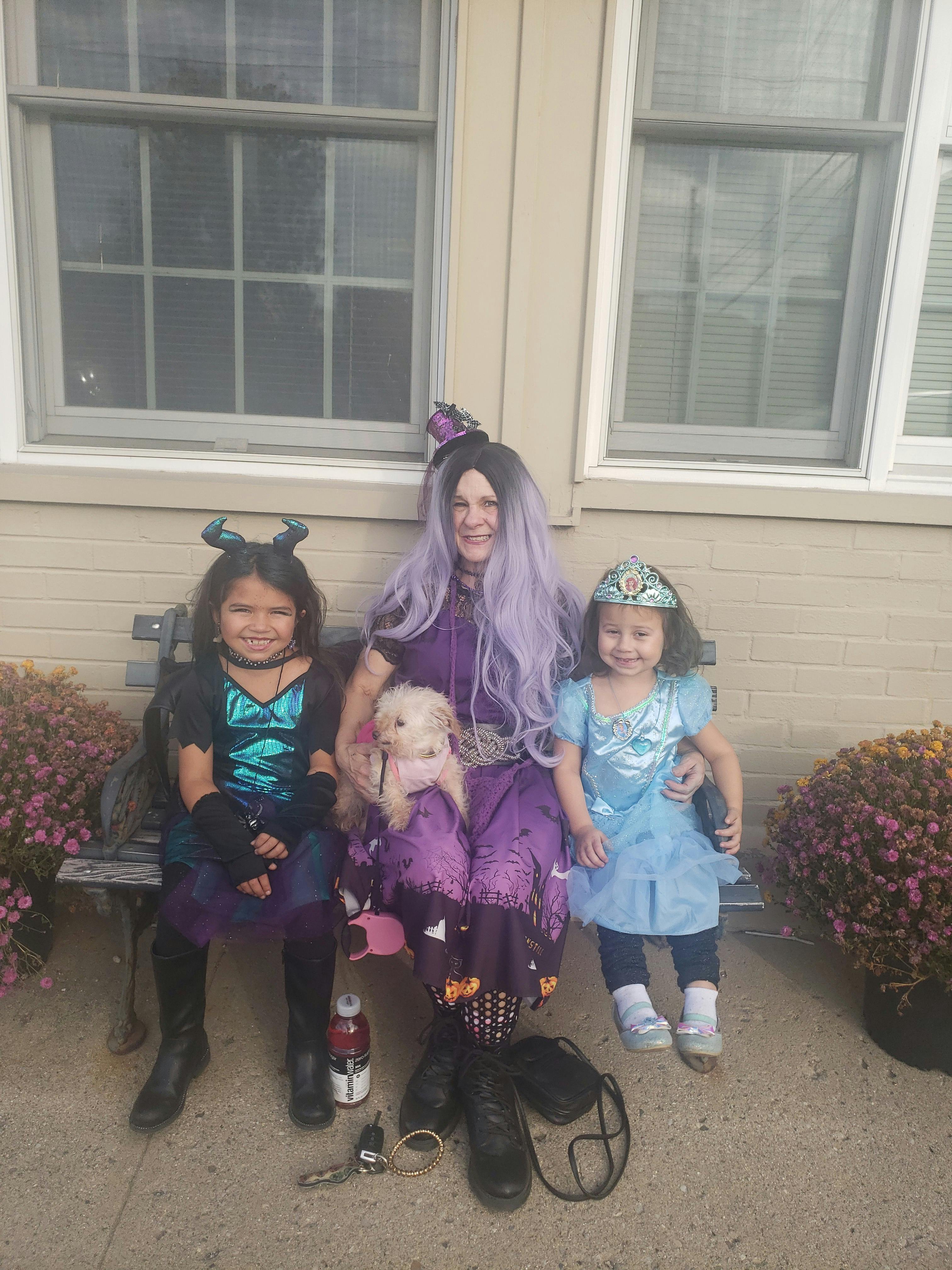 Person in purple wig and two children in costumes sitting on a bench