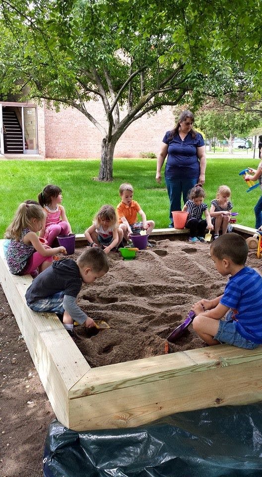 Children playing in a sandbox with a person standing nearby