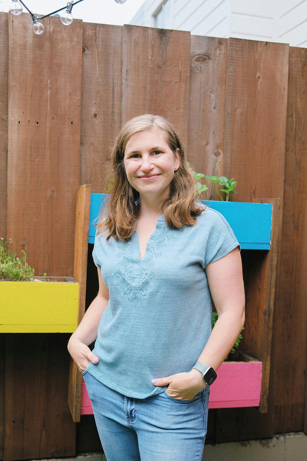 A person standing with hands in pockets in front of a wooden fence with planters