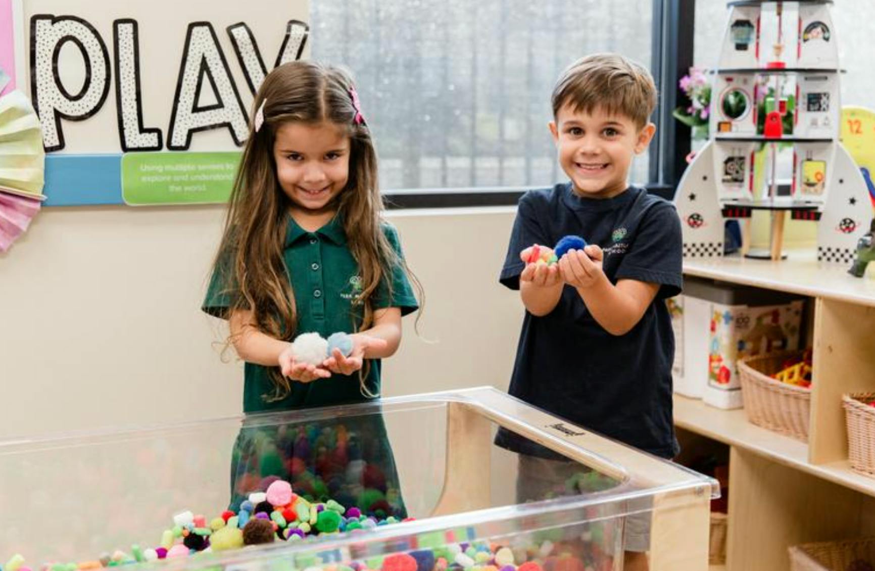 Two children holding colorful pom-poms over a sensory bin in a classroom