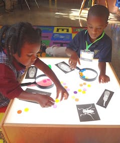 Two children playing with colorful shapes and magnifying glasses on a light table