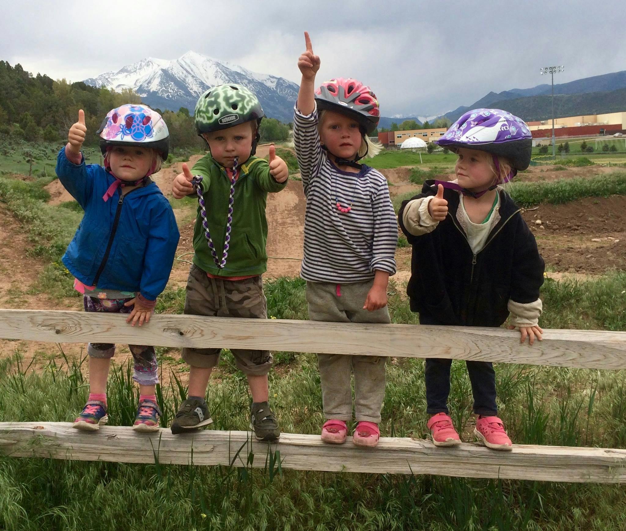Four children in helmets standing on a wooden fence, giving thumbs up and pointing