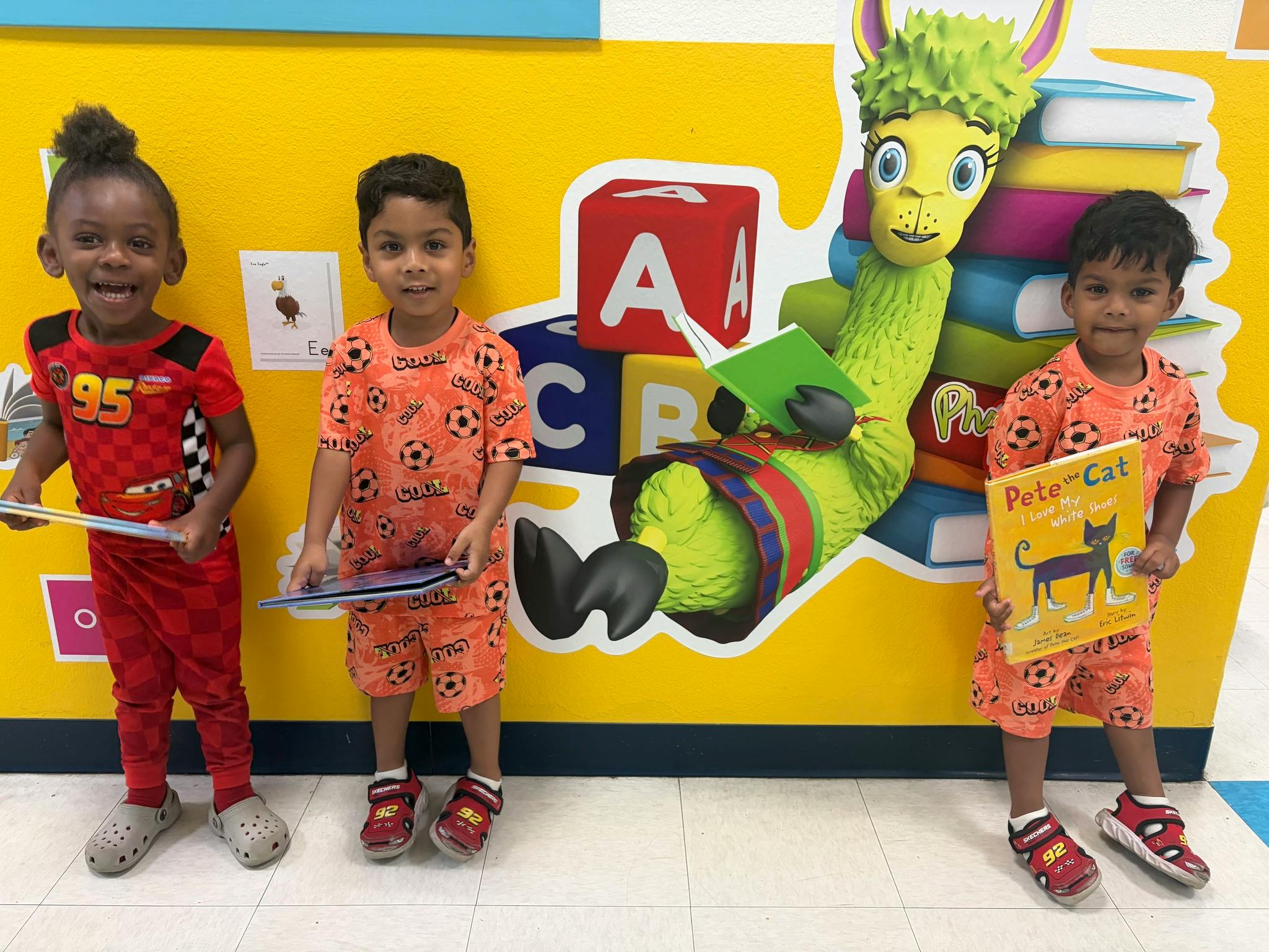 Three children holding books in front of a colorful wall mural with a character