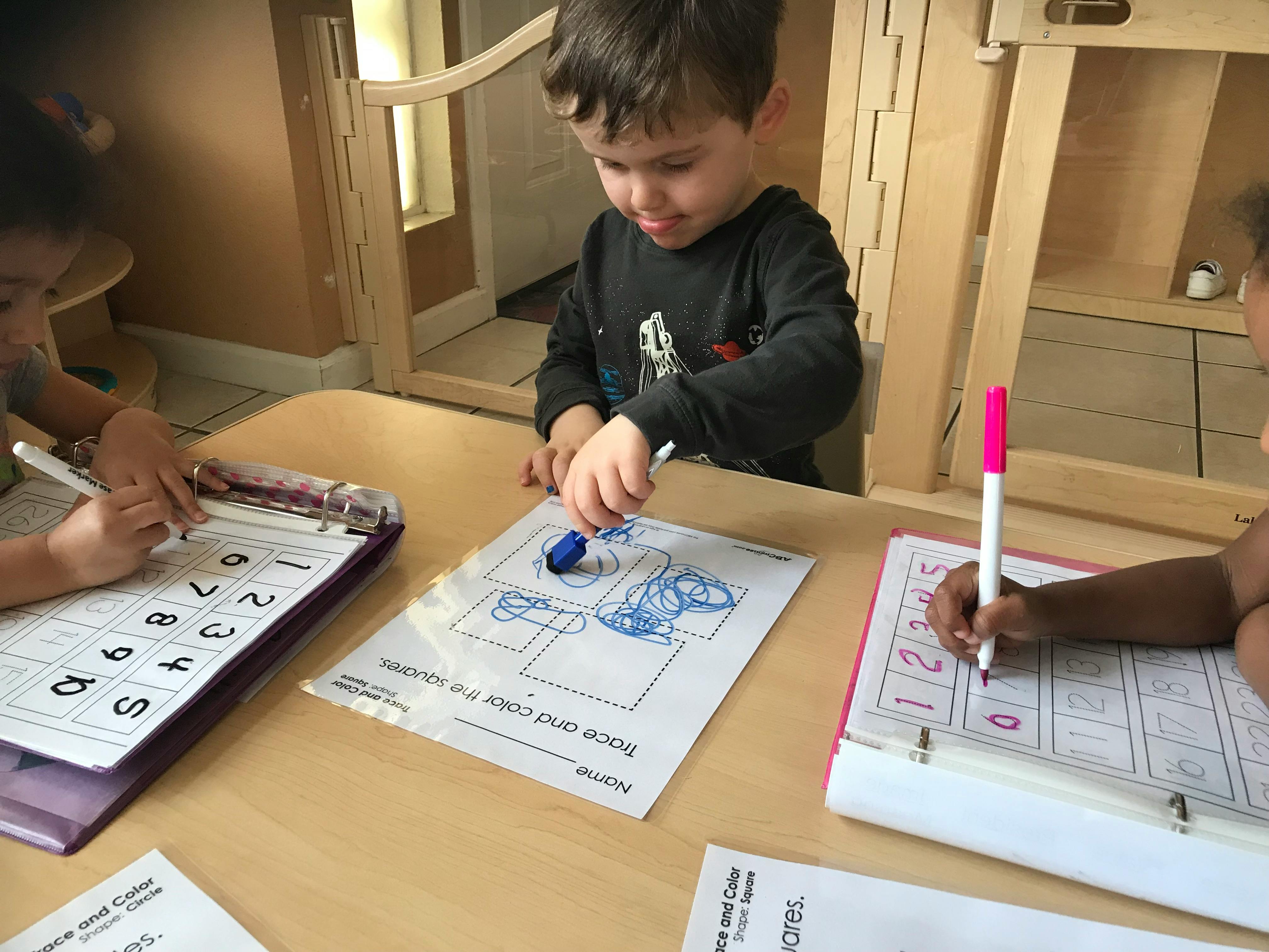 Child coloring a drawing at a table with other children writing