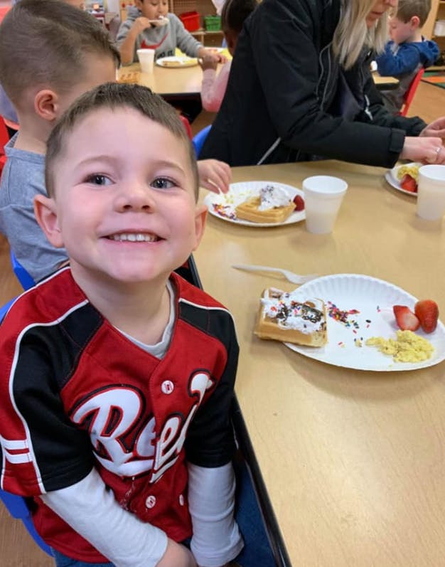Child in a red and black shirt smiling at a table with food