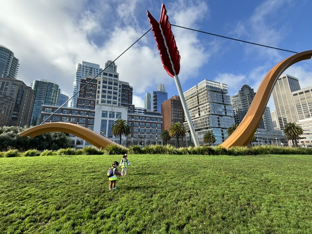 Two children on a grassy hill with a large bow and arrow sculpture and city buildings