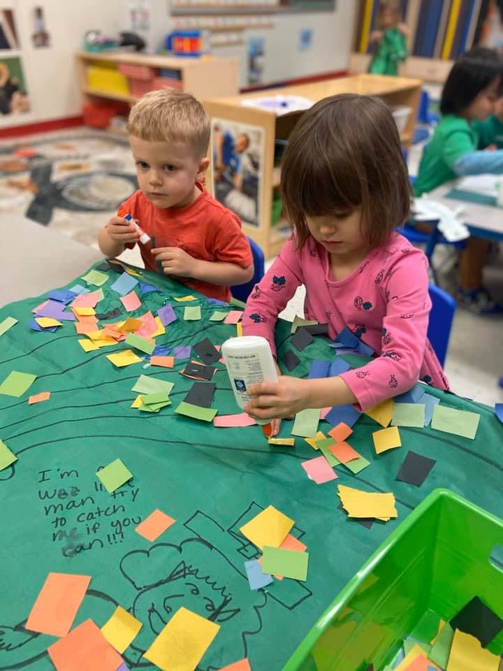 Two children gluing paper squares onto a green surface in a classroom