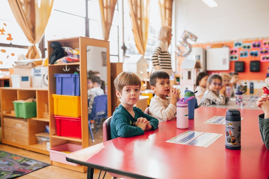 Children sitting at a red table in a classroom with a person standing in the background