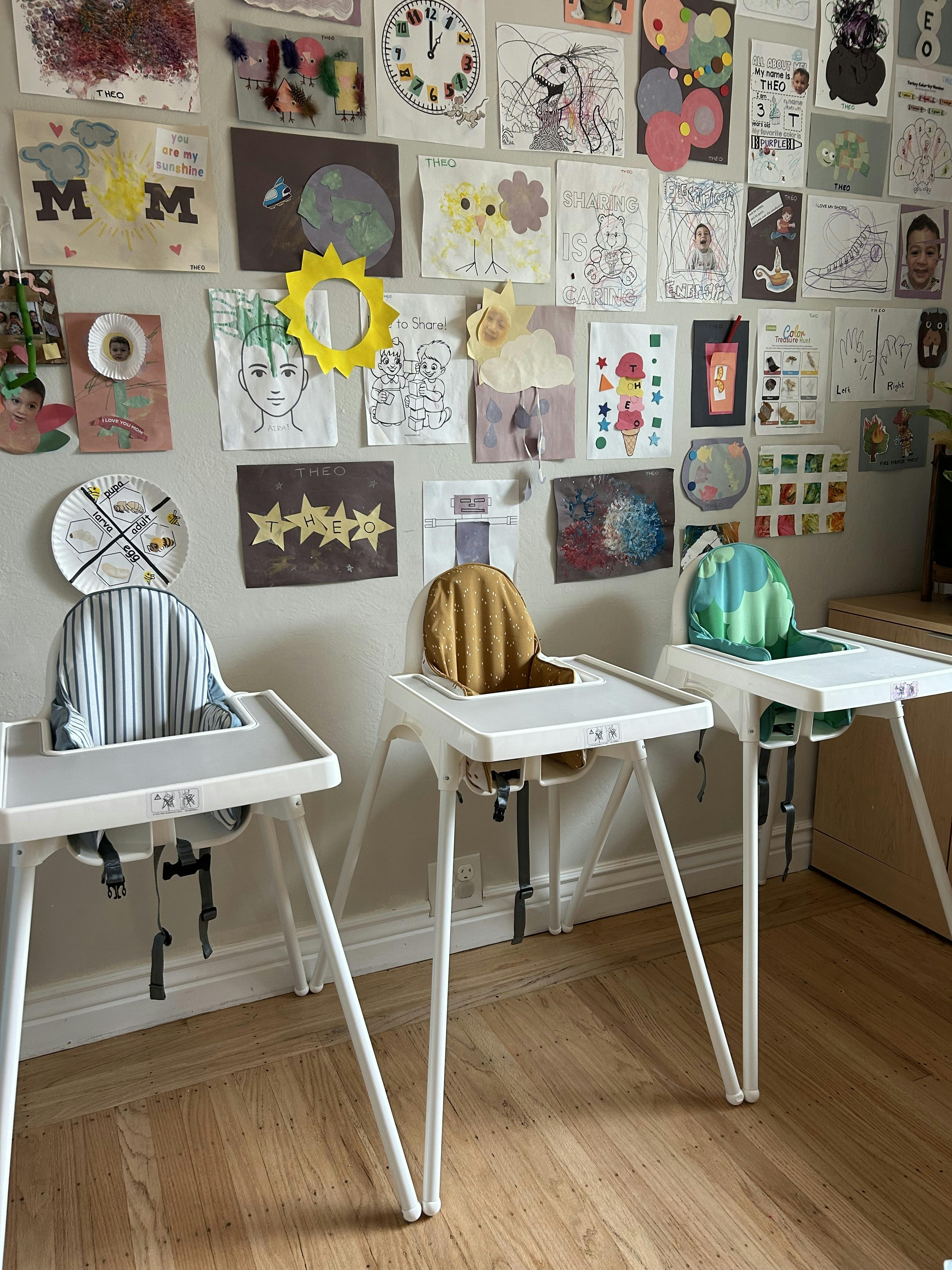 Three empty high chairs lined up against a wall covered in children's art