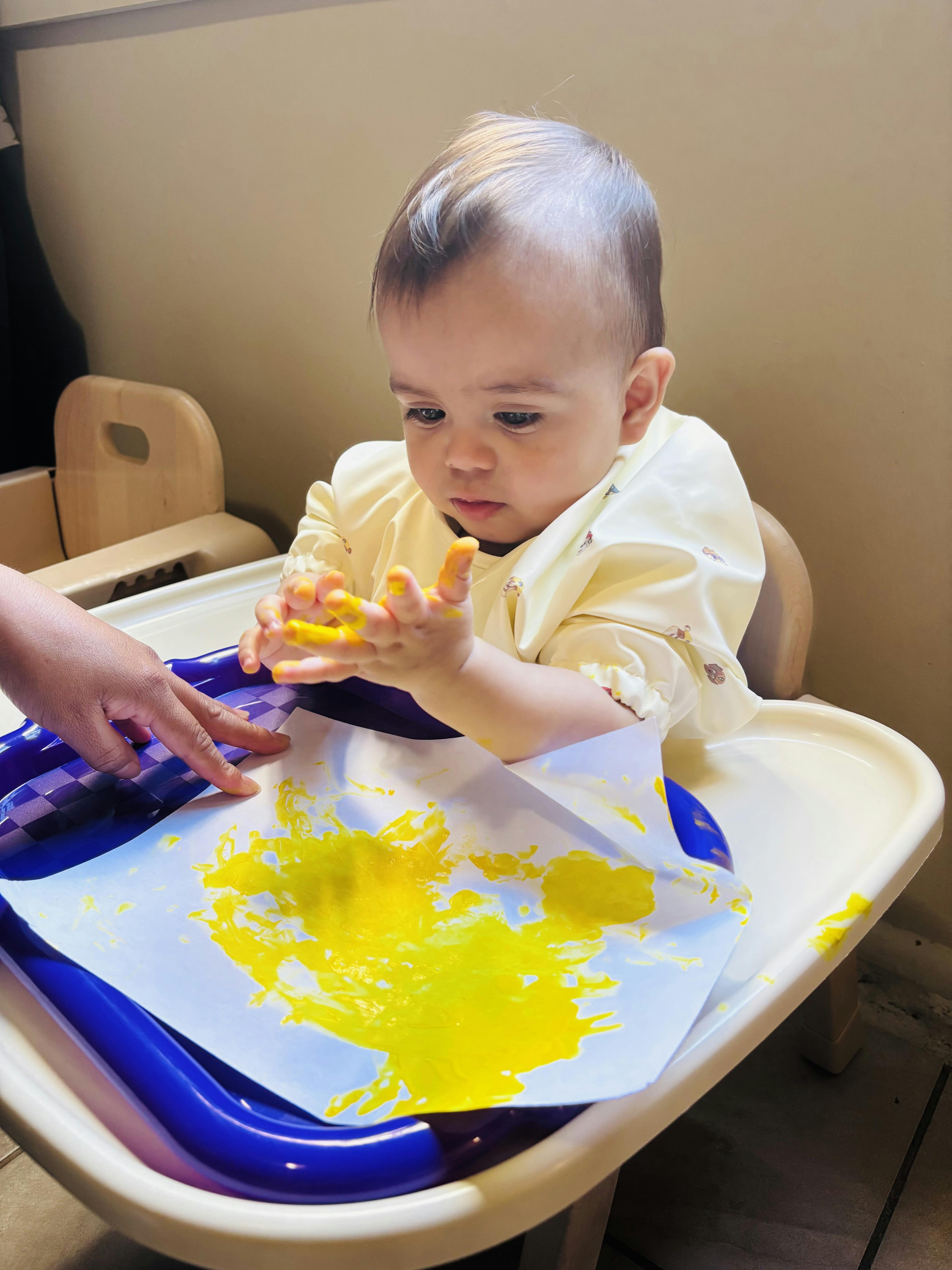 Child finger painting with yellow paint on paper in a high chair