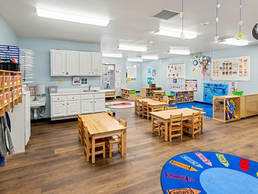 Classroom with small wooden tables and chairs, a sink area, and colorful wall decorations