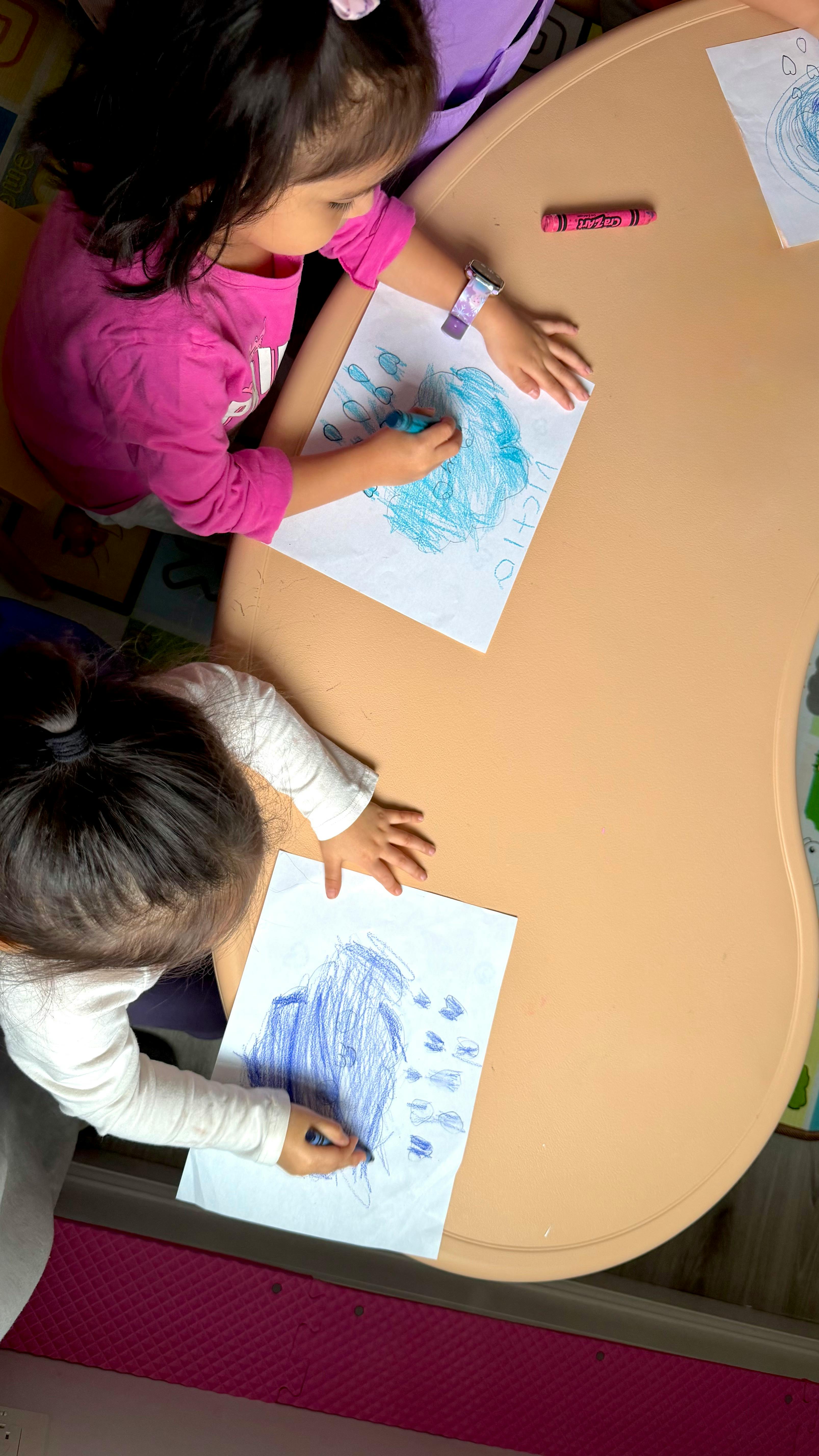 Two children sitting at a table and drawing with crayons on paper