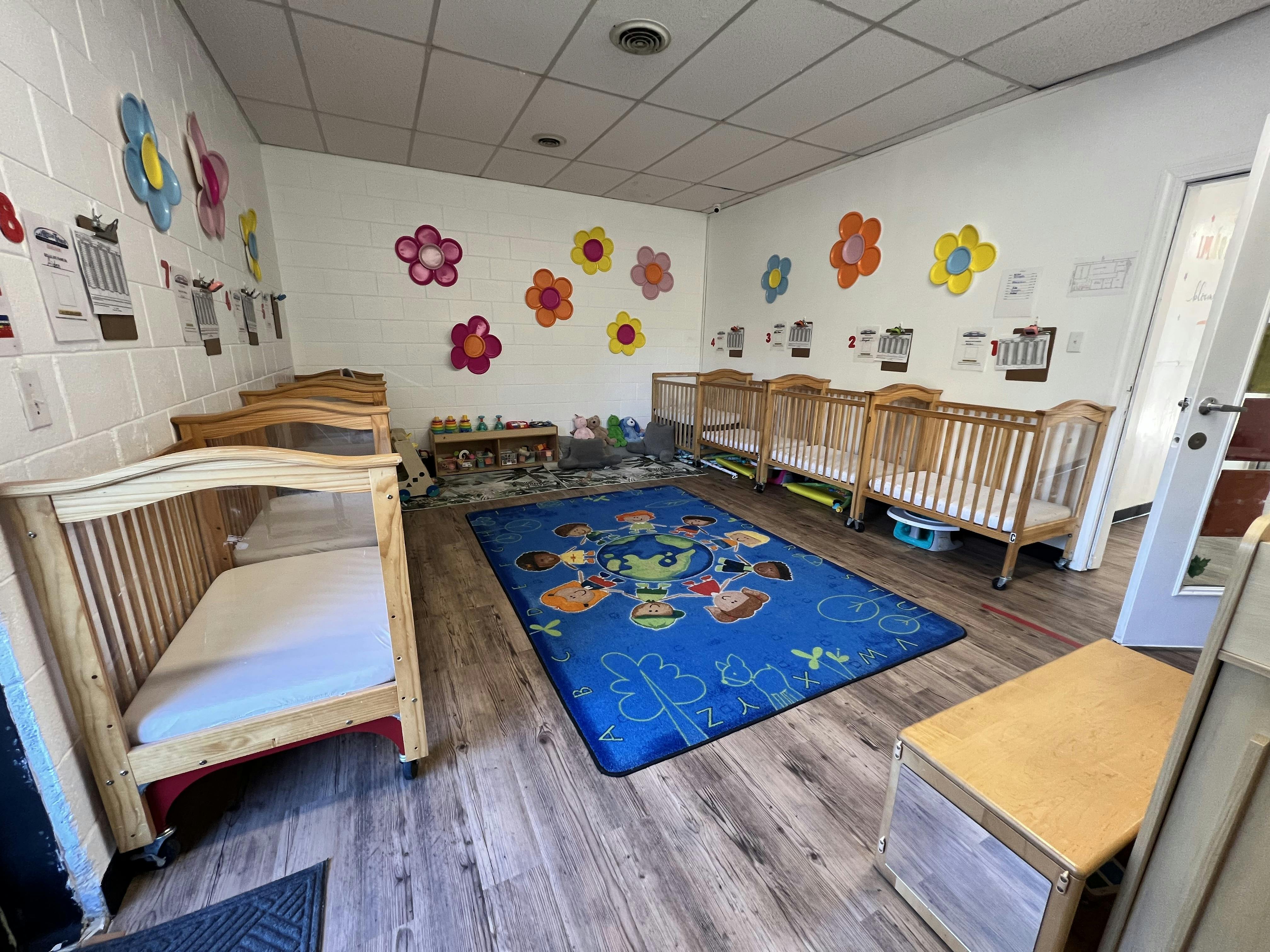 A room with multiple wooden cribs, a blue rug with children on it, and toy shelves