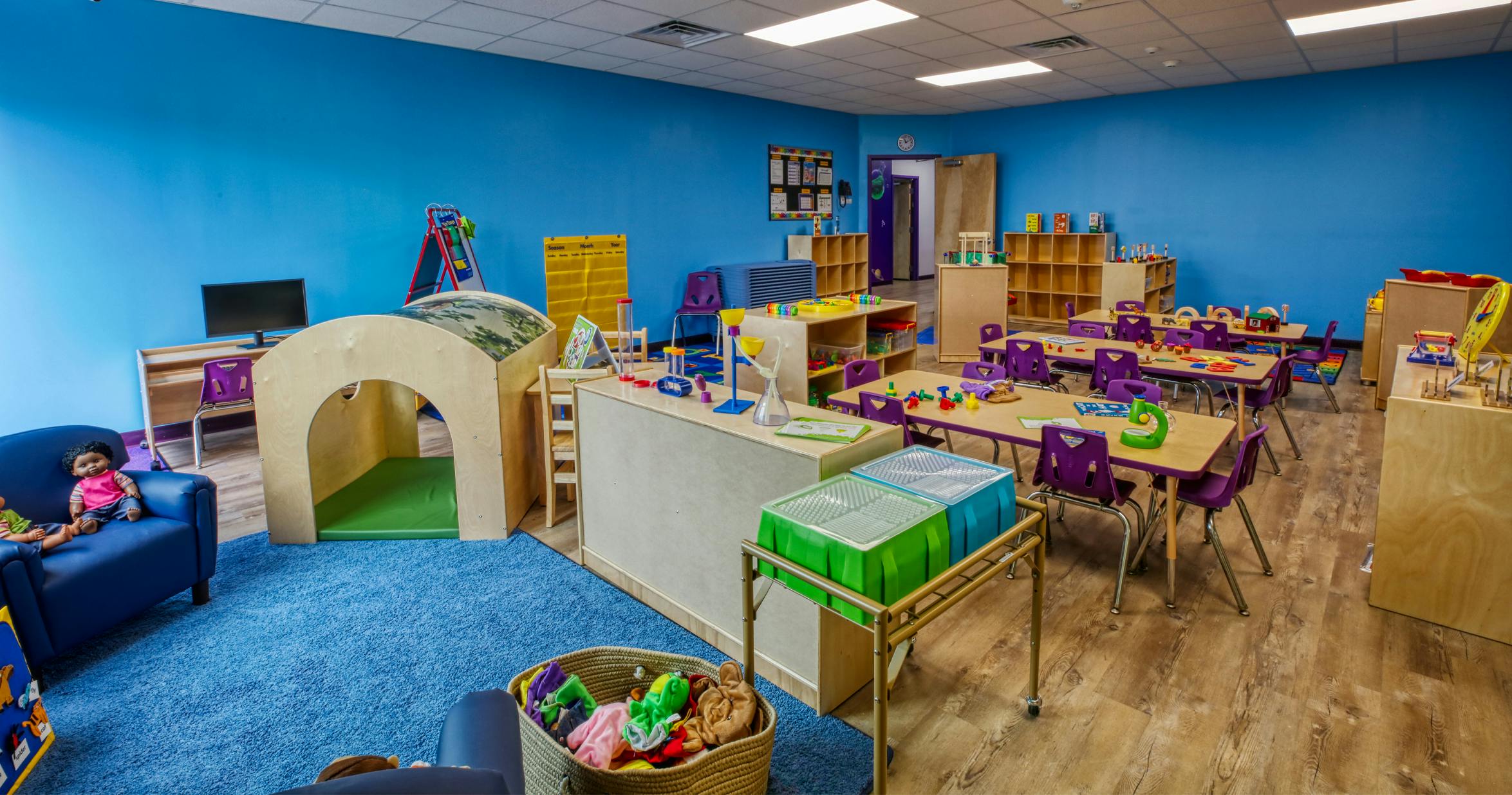 Classroom with blue walls, small tables with purple chairs, and play equipment