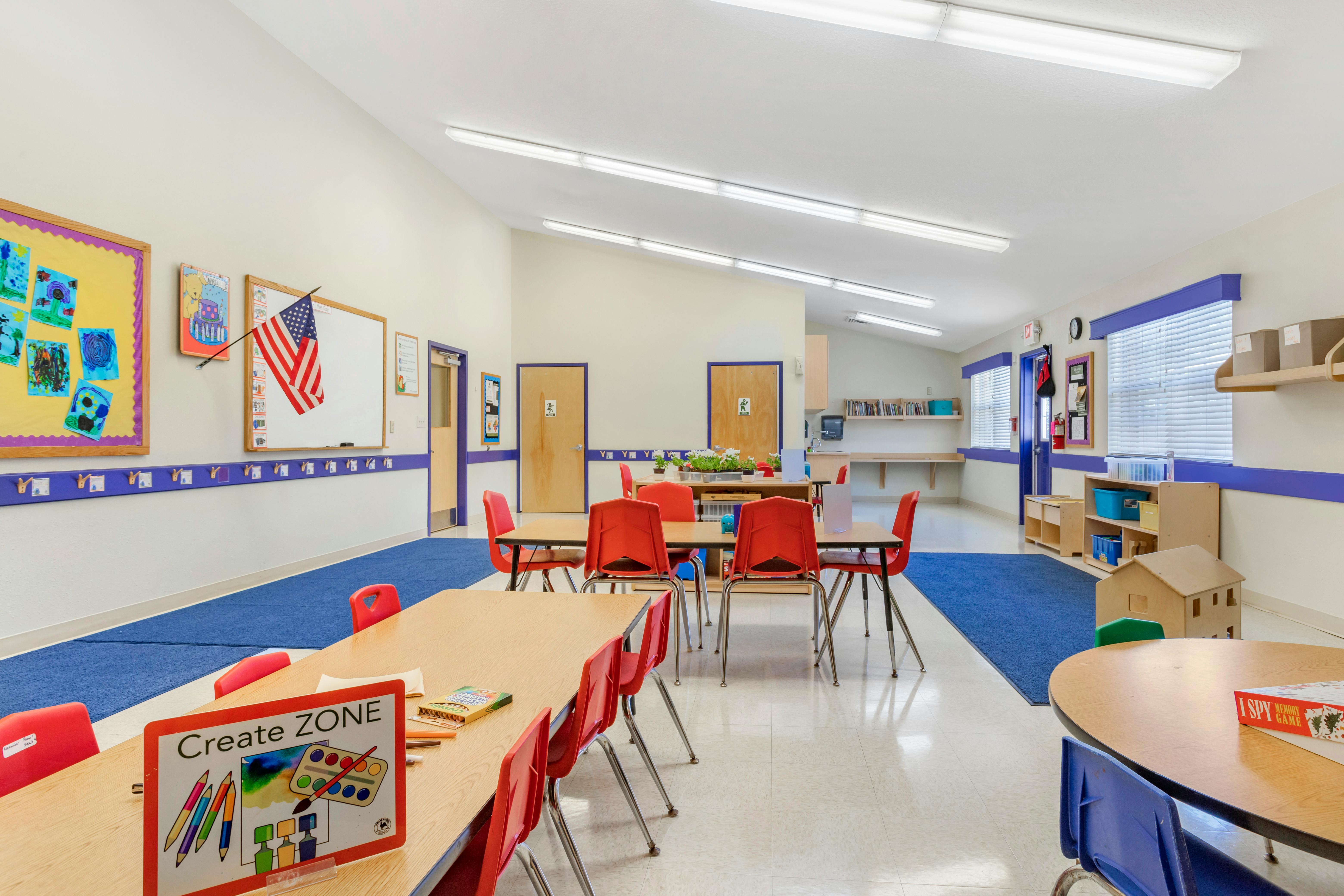 Classroom with child-sized tables and chairs, American flag on whiteboard, and artwork