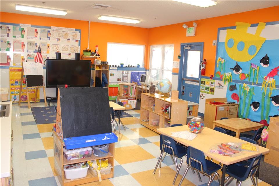 A colorful classroom with small tables and chairs, a television, and a chalkboard