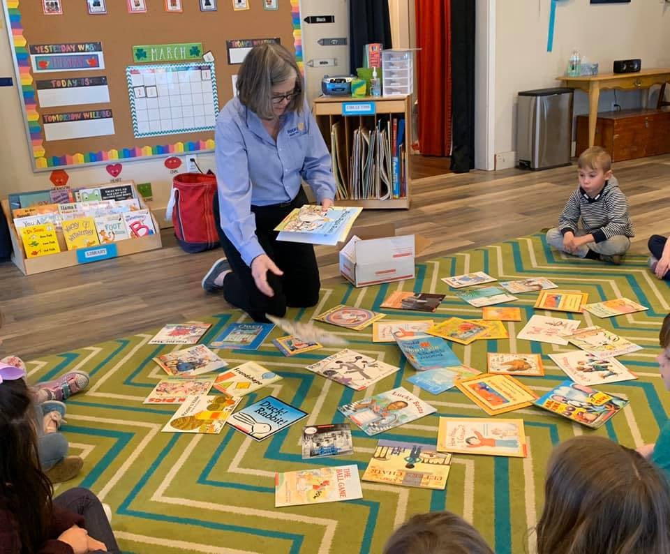 Person kneeling on a rug, holding a book, surrounded by books and children