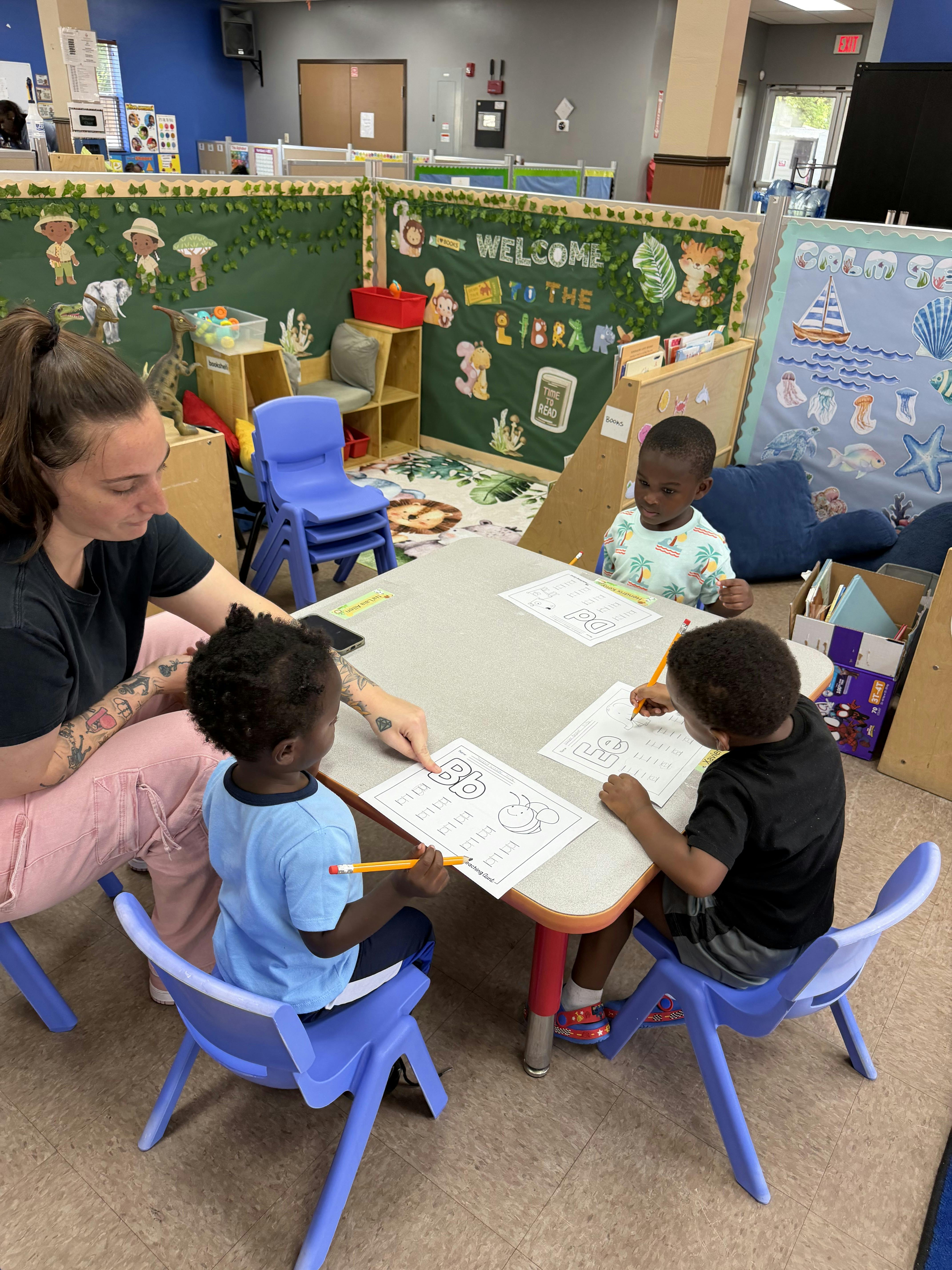 Teacher pointing to a worksheet for three children practicing writing at a table