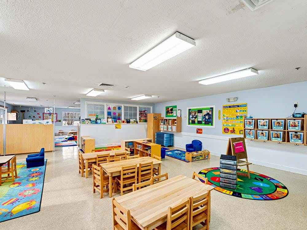Empty classroom with small wooden tables and chairs, colorful rugs, and wall decorations