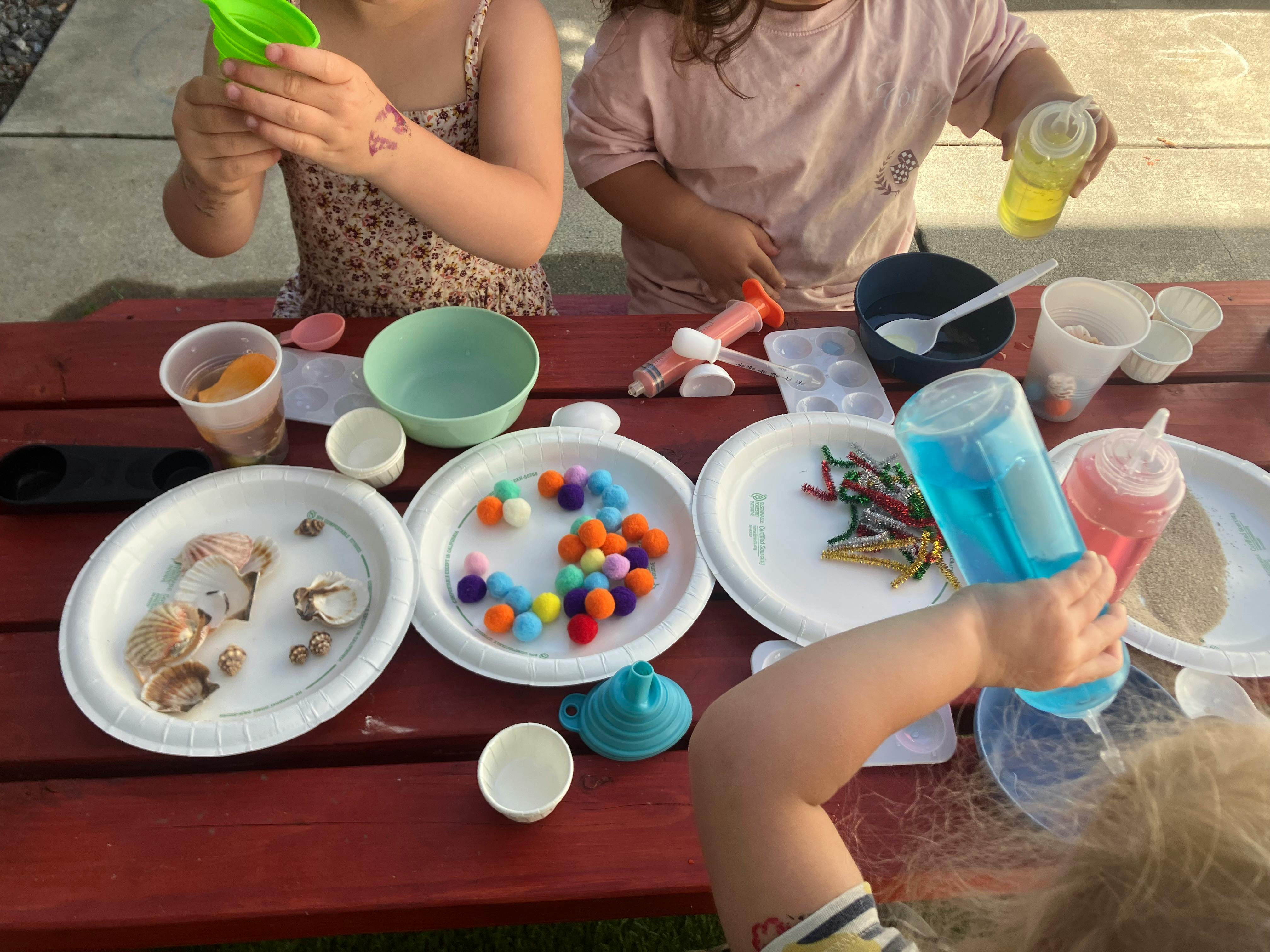 Children playing with liquids, colorful pom poms, and shells at an outdoor table