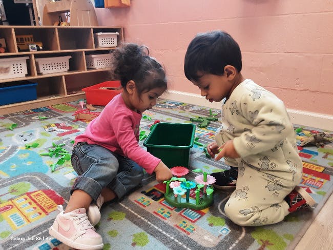 Two children playing with toy flowers and a green bucket on a rug