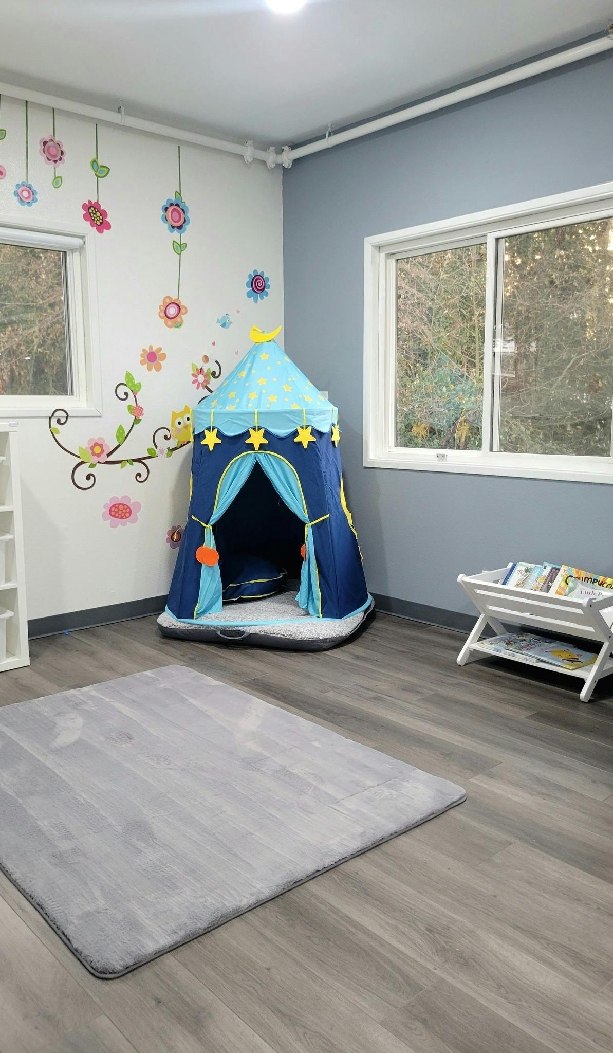 A room with a blue play tent, a white bookshelf with books, and a gray rug
