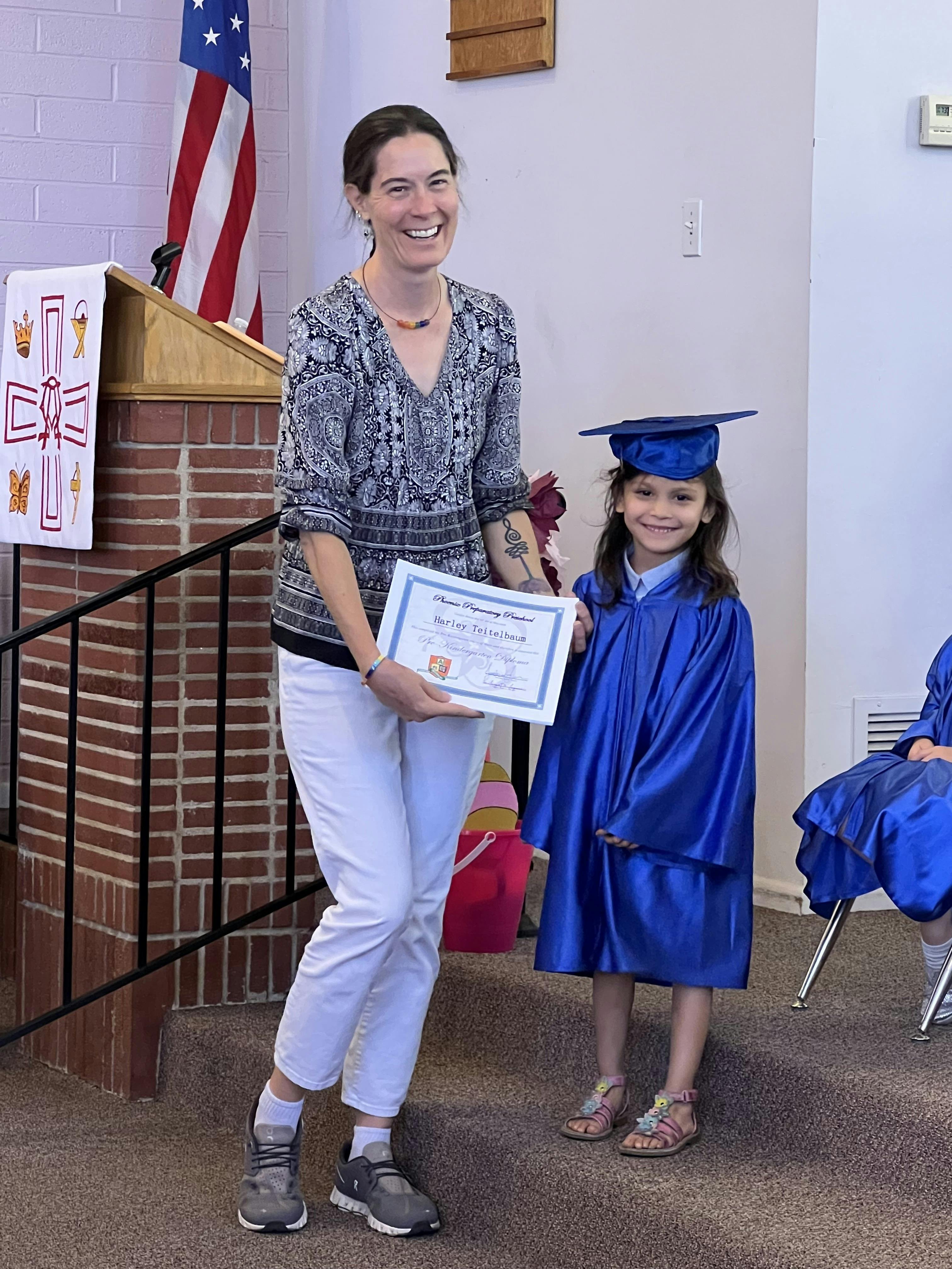 Adult handing a preschool graduation certificate to a child in a cap and gown