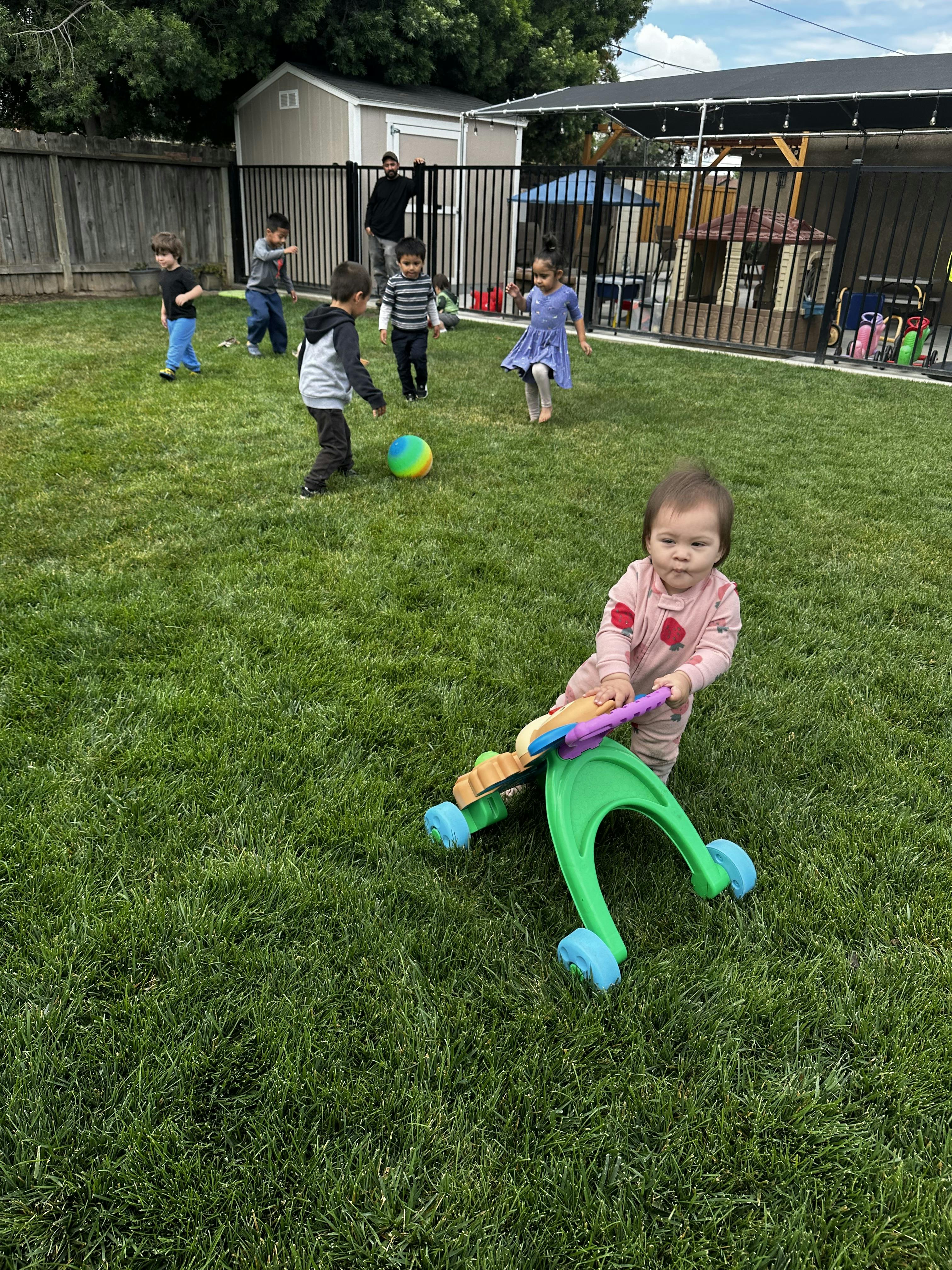 Child pushing a toy walker on grass, other children playing with a ball in background