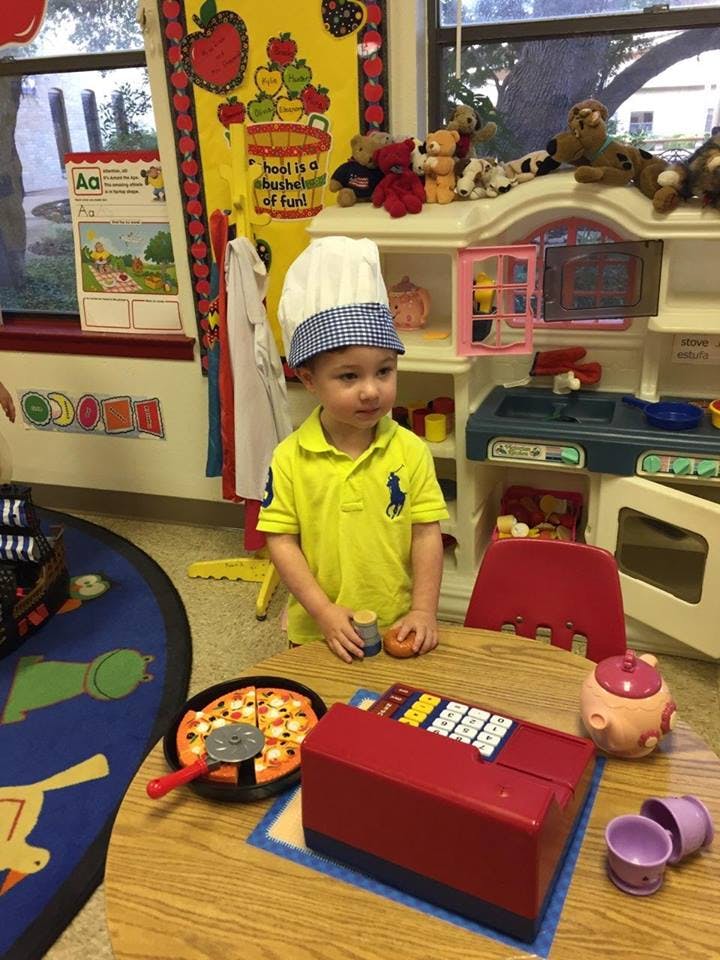 Child in a chef hat standing at a table with toy pizza and cash register