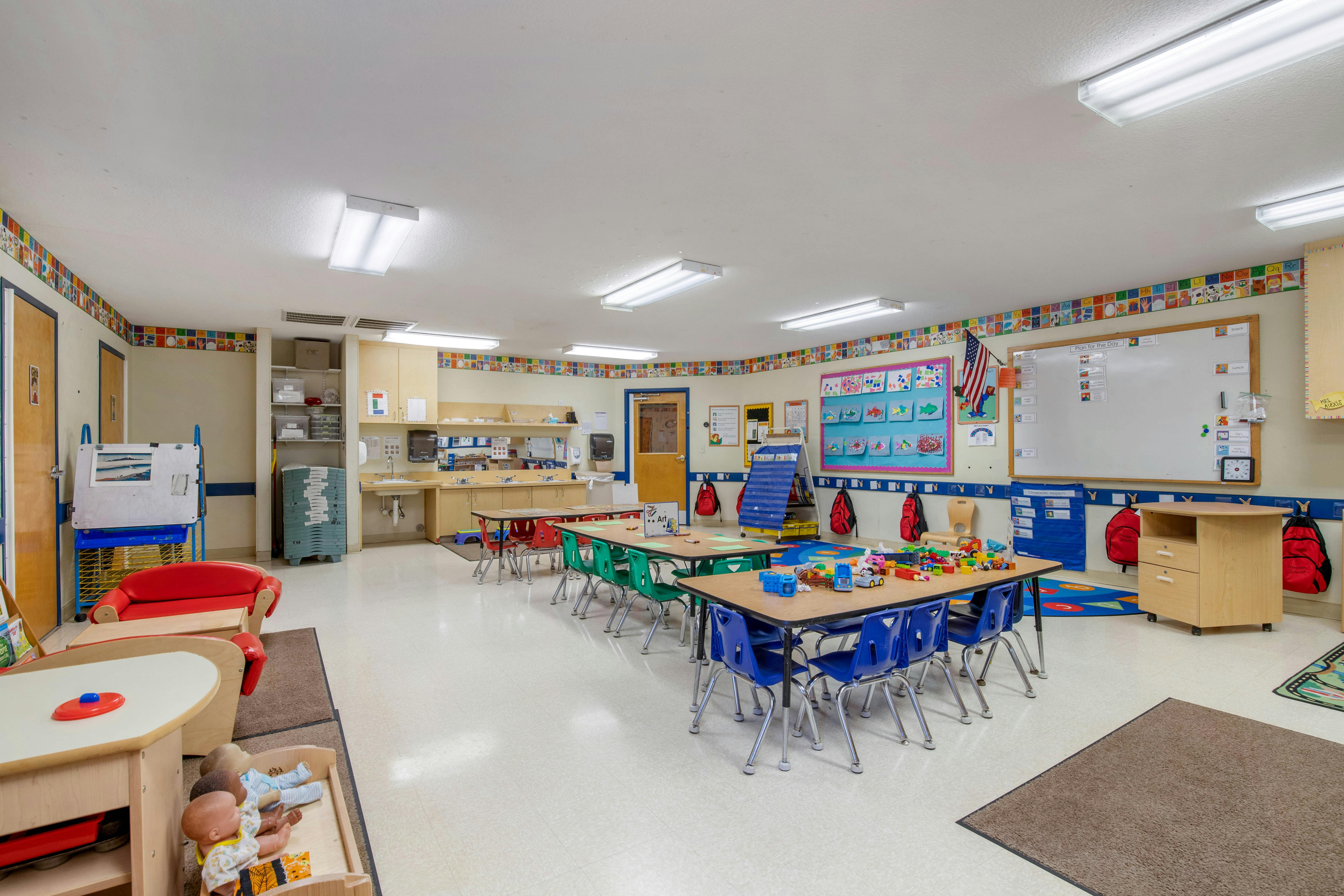 An empty classroom with small tables and chairs, toys, and learning materials